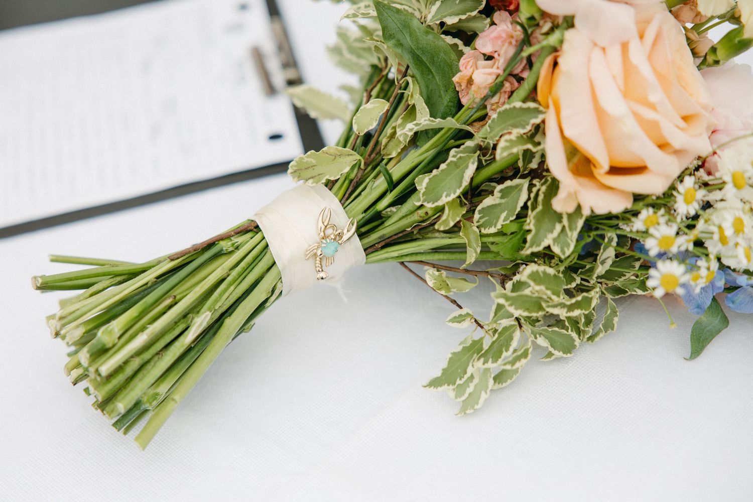 Elegant bridal bouquet with peach roses, daisies, and greenery resting on white surface beside clipboard.