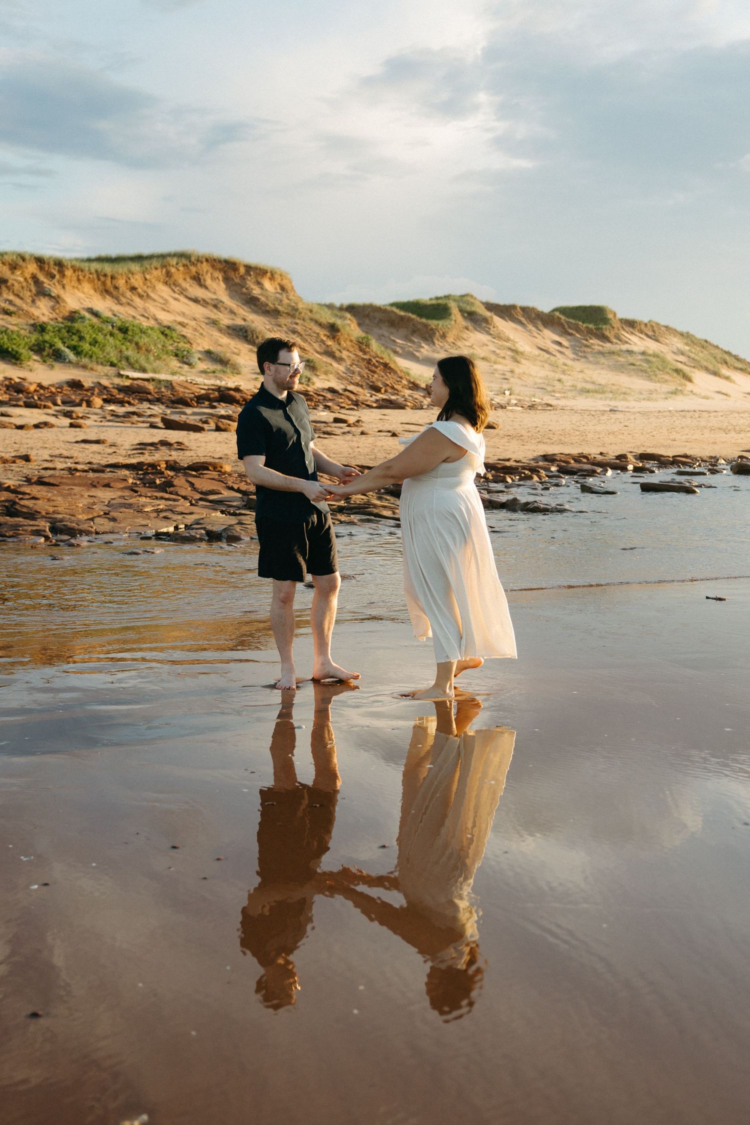 Couple holding hands on a reflective wet beach with sandy dunes in the background.