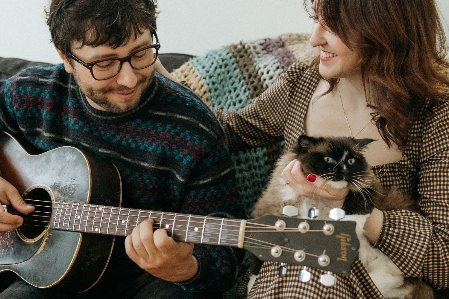 Man playing acoustic guitar on sofa while smiling woman holds a Ragdoll cat in cozy home setting.