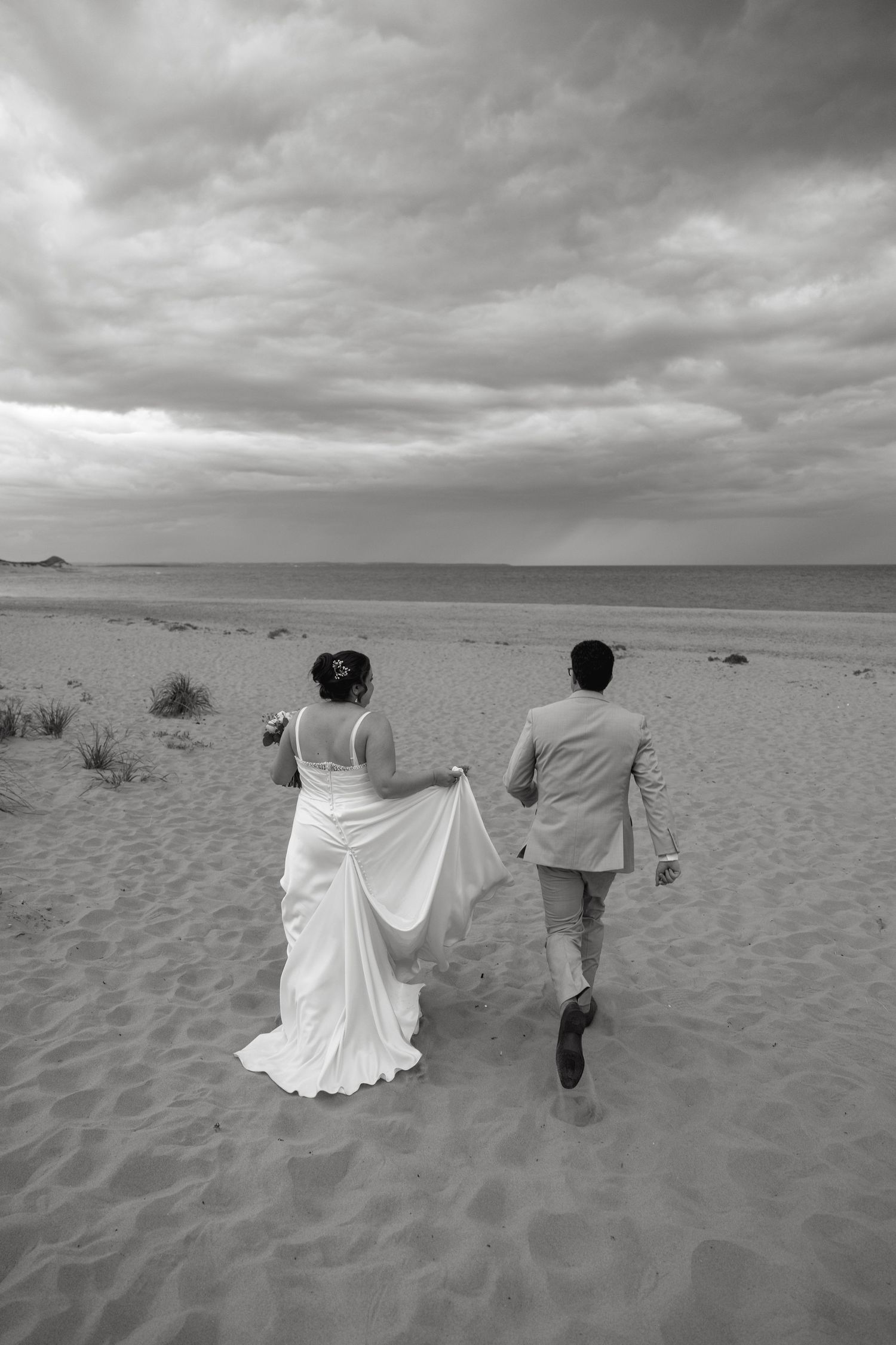 Black and white photo of bride and groom walking on sandy beach.