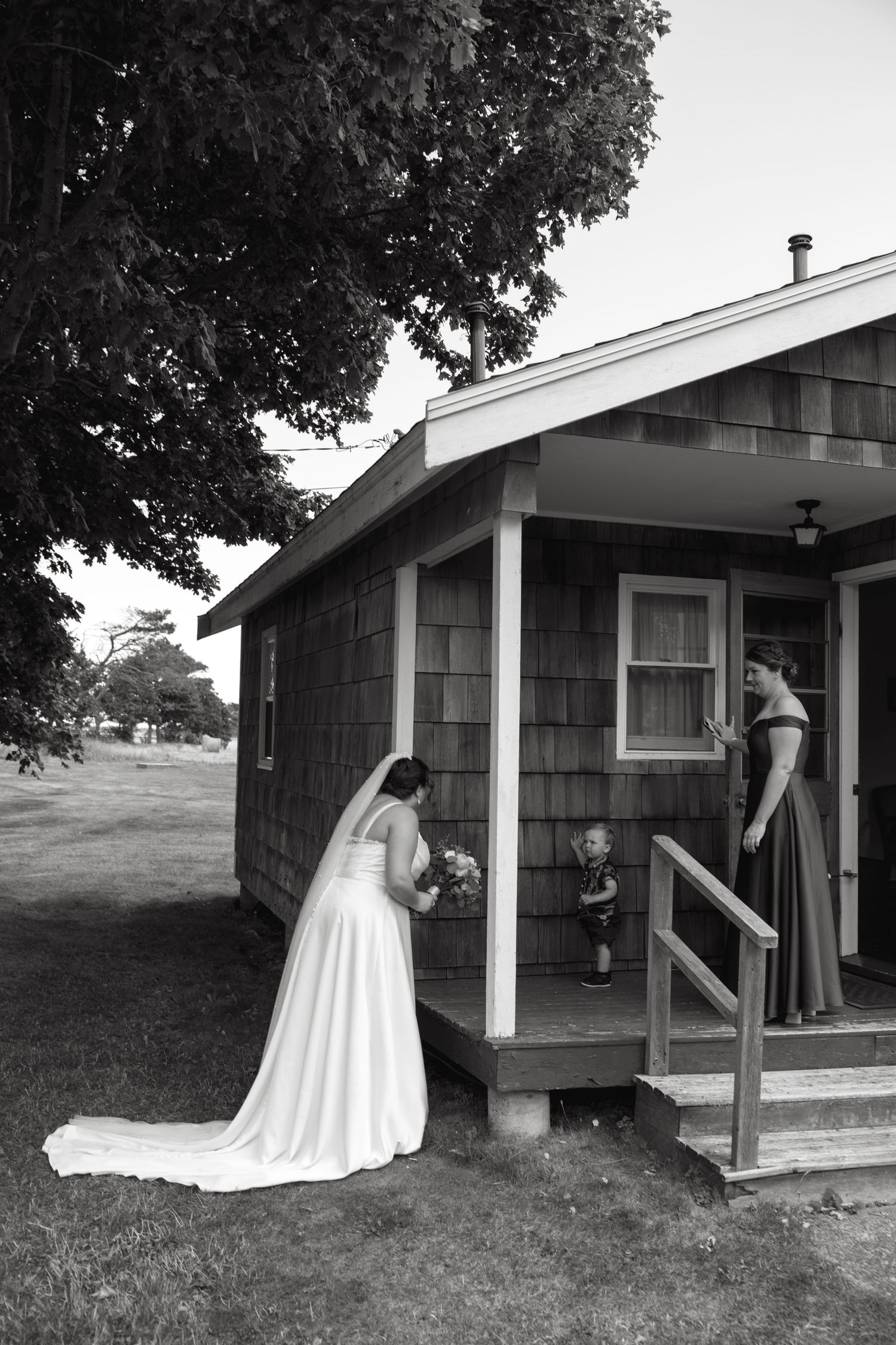 Bride in white gown holds bouquet outside rustic cottage while bridesmaid and child watch from porch steps.
