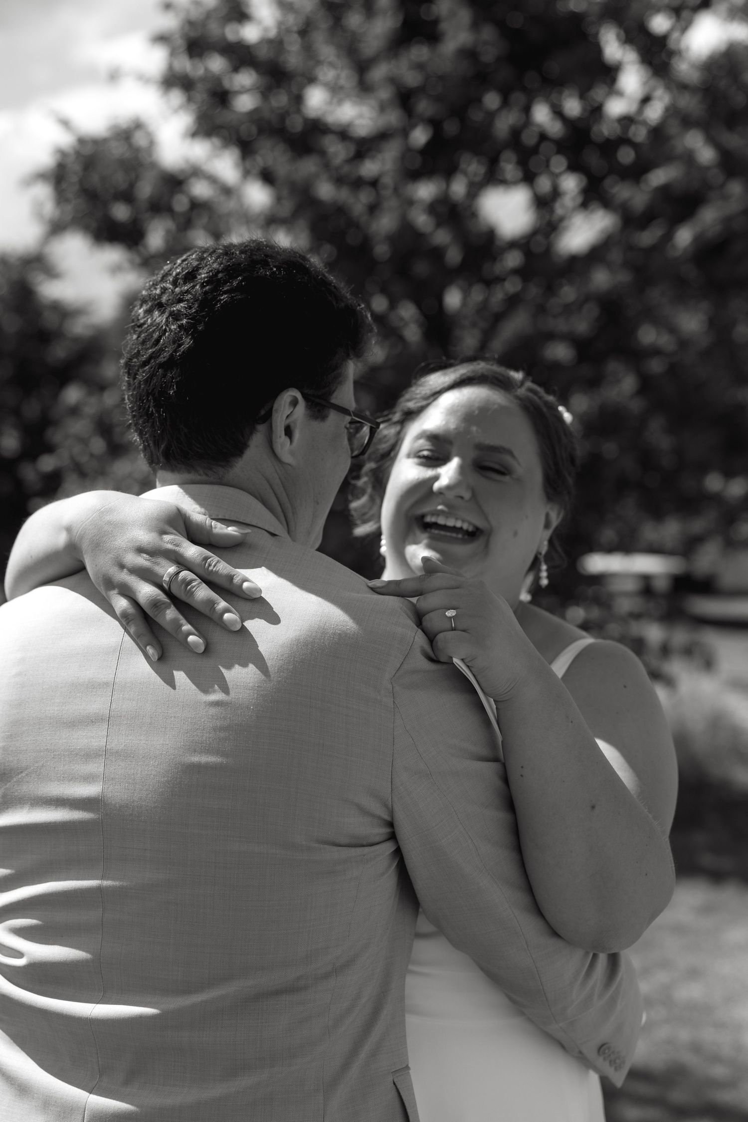 Black and white photo of a laughing bride embracing her groom outdoors at a wedding celebration.