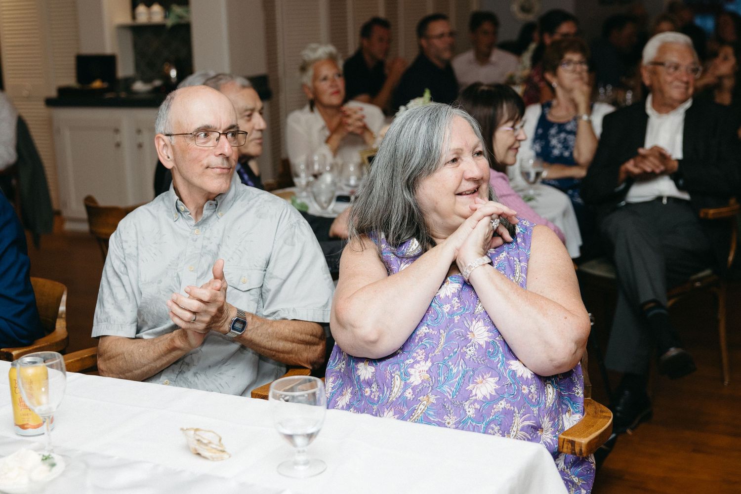 Older couple smiles and claps joyfully while seated at wedding reception dinner table.