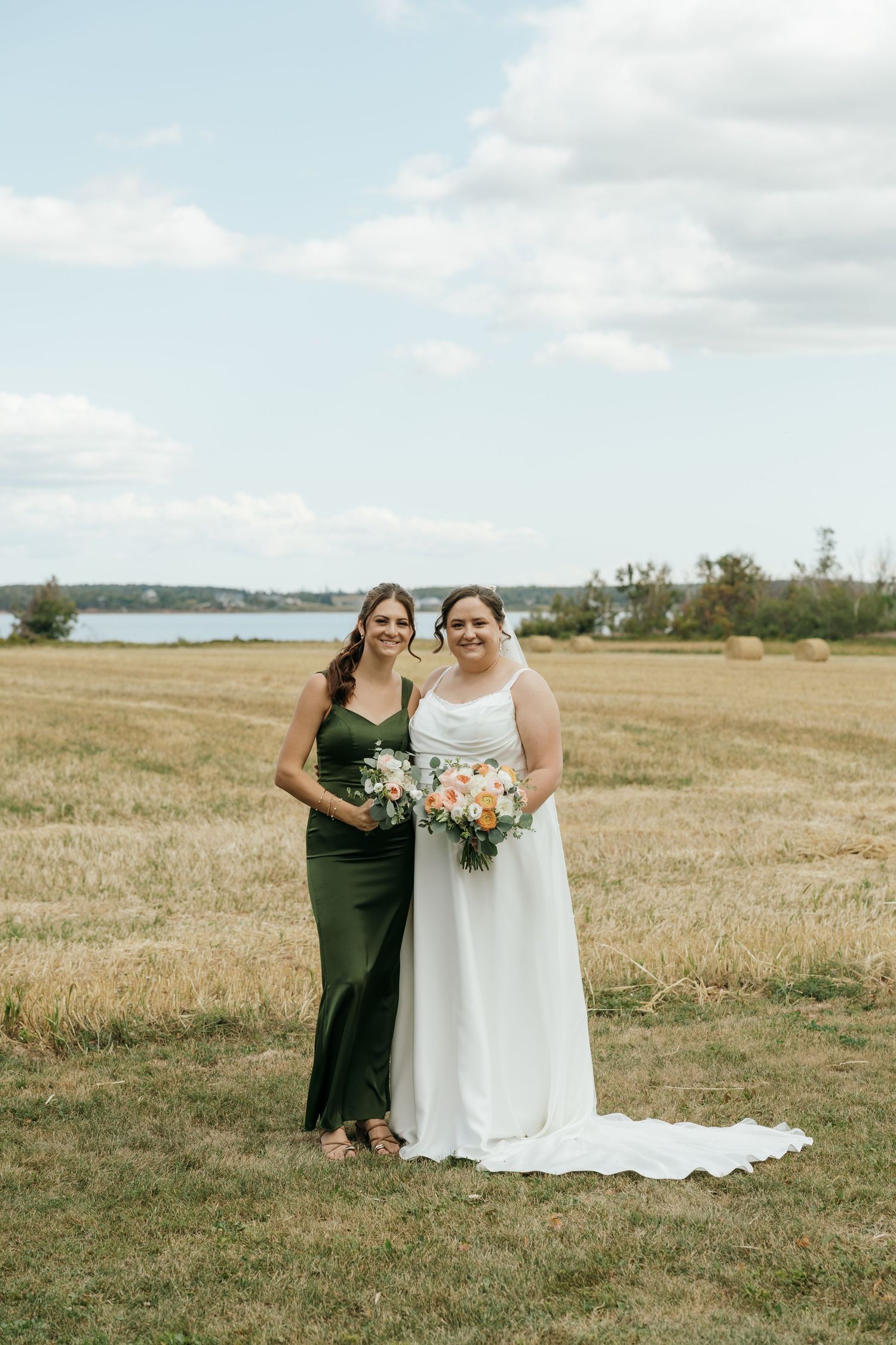 Bride in white gown and bridesmaid in olive green dress posing in a harvested field by a lake.