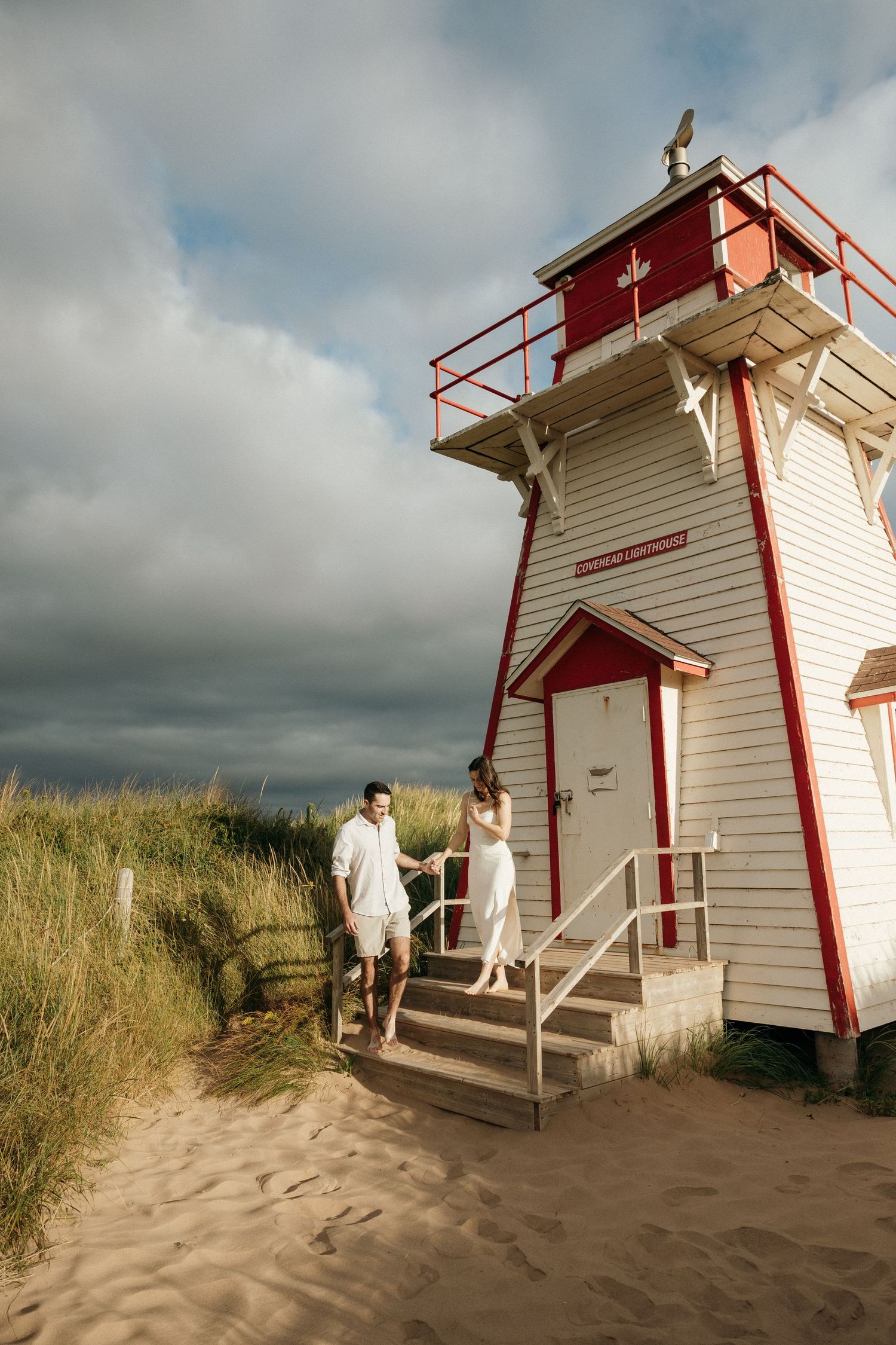 Red and white lighthouse on sandy beach with dramatic clouds and tall grass.