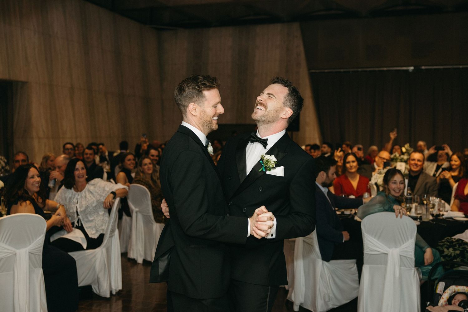 Two people dancing and laughing together at an elegant wedding reception with guests in the background.
