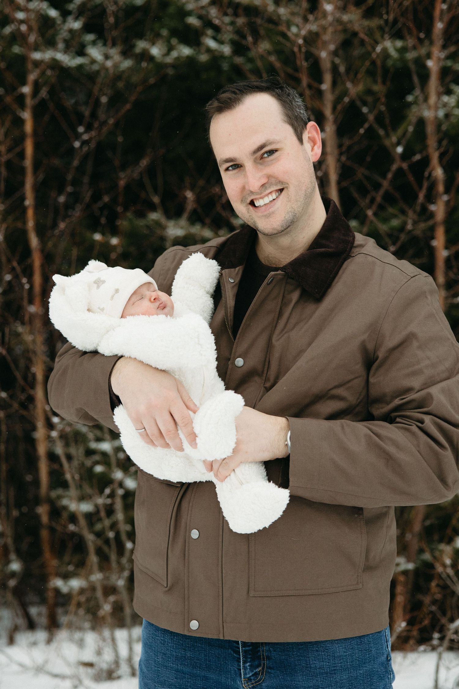 A person in a brown jacket holds a baby wrapped in white against a snowy winter background.