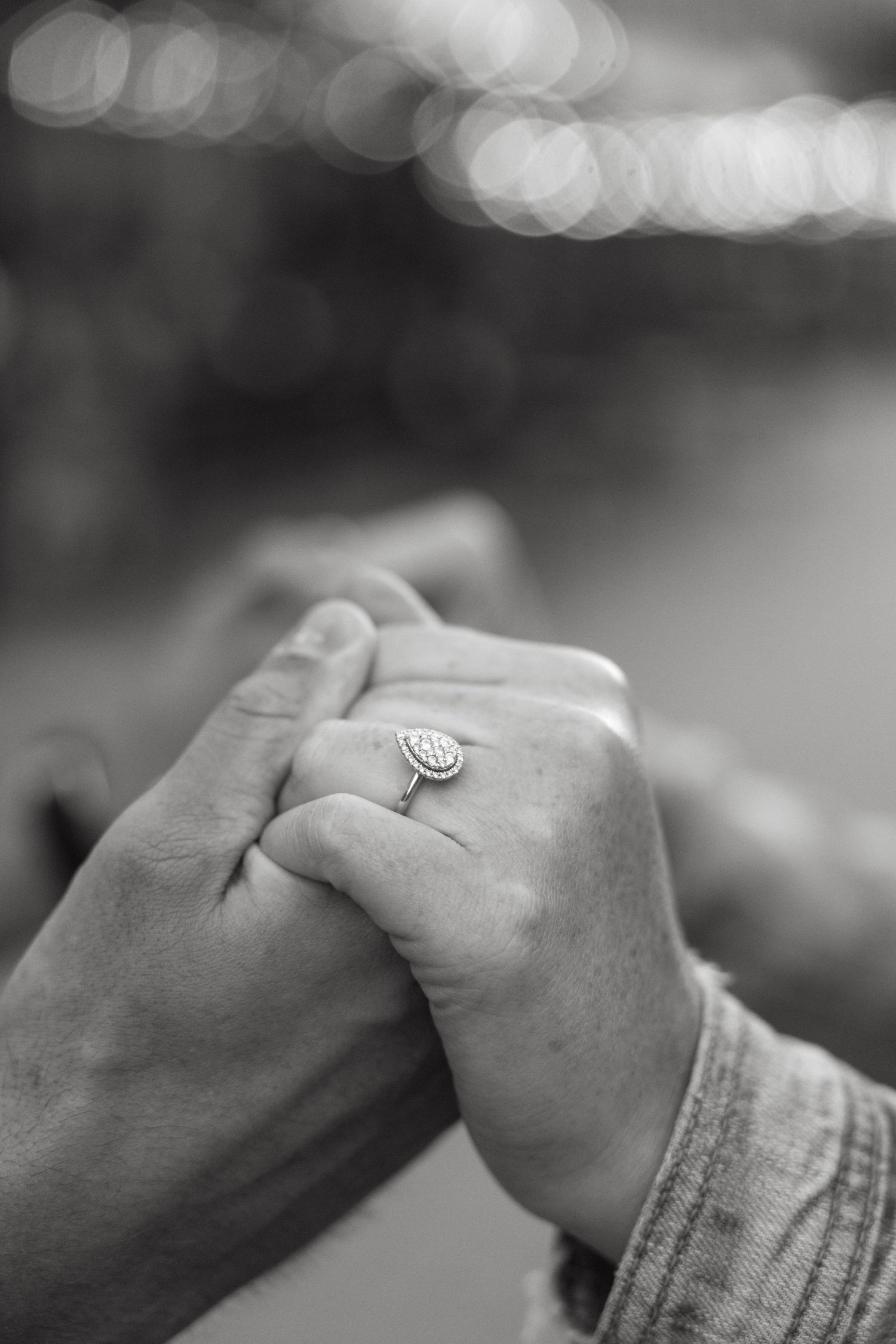 A close-up black and white photo of clasped hands with an engagement ring.