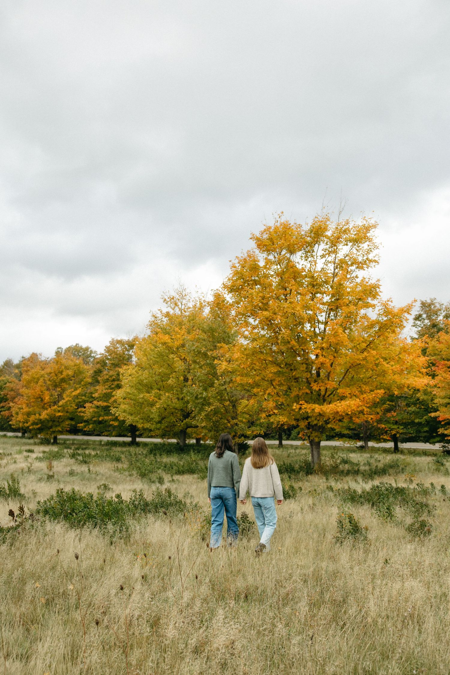 Two women walk hand in hand through a grassy field toward vibrant orange and yellow autumn trees under a cloudy sky.