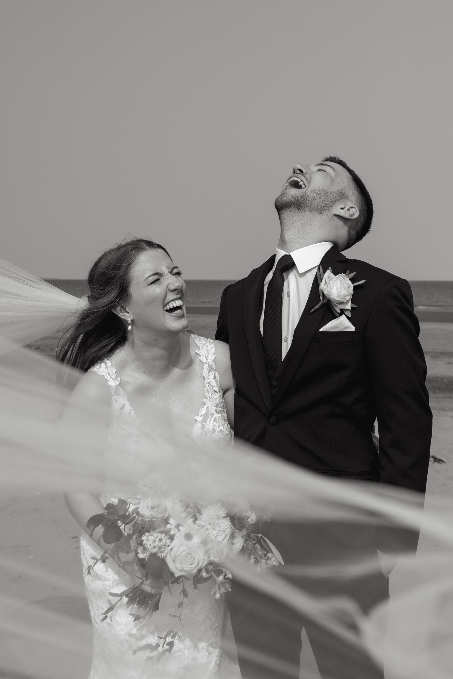 Joyful wedding couple laughing together as veil flows in the wind on beach.