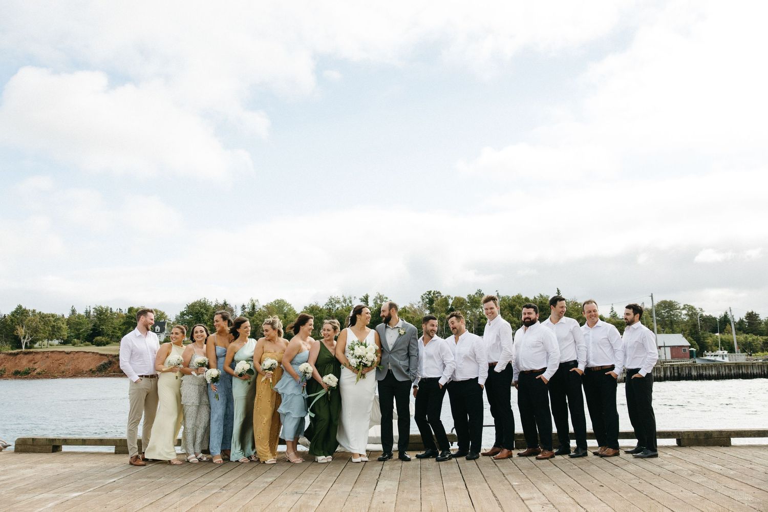Large wedding party lined up for panoramic photo by waterfront.