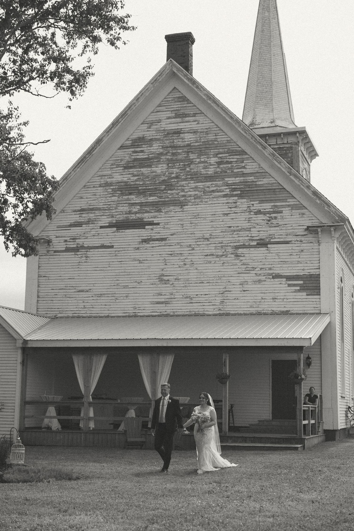 Black and white photo of couple walking outside rustic white church with steeple.