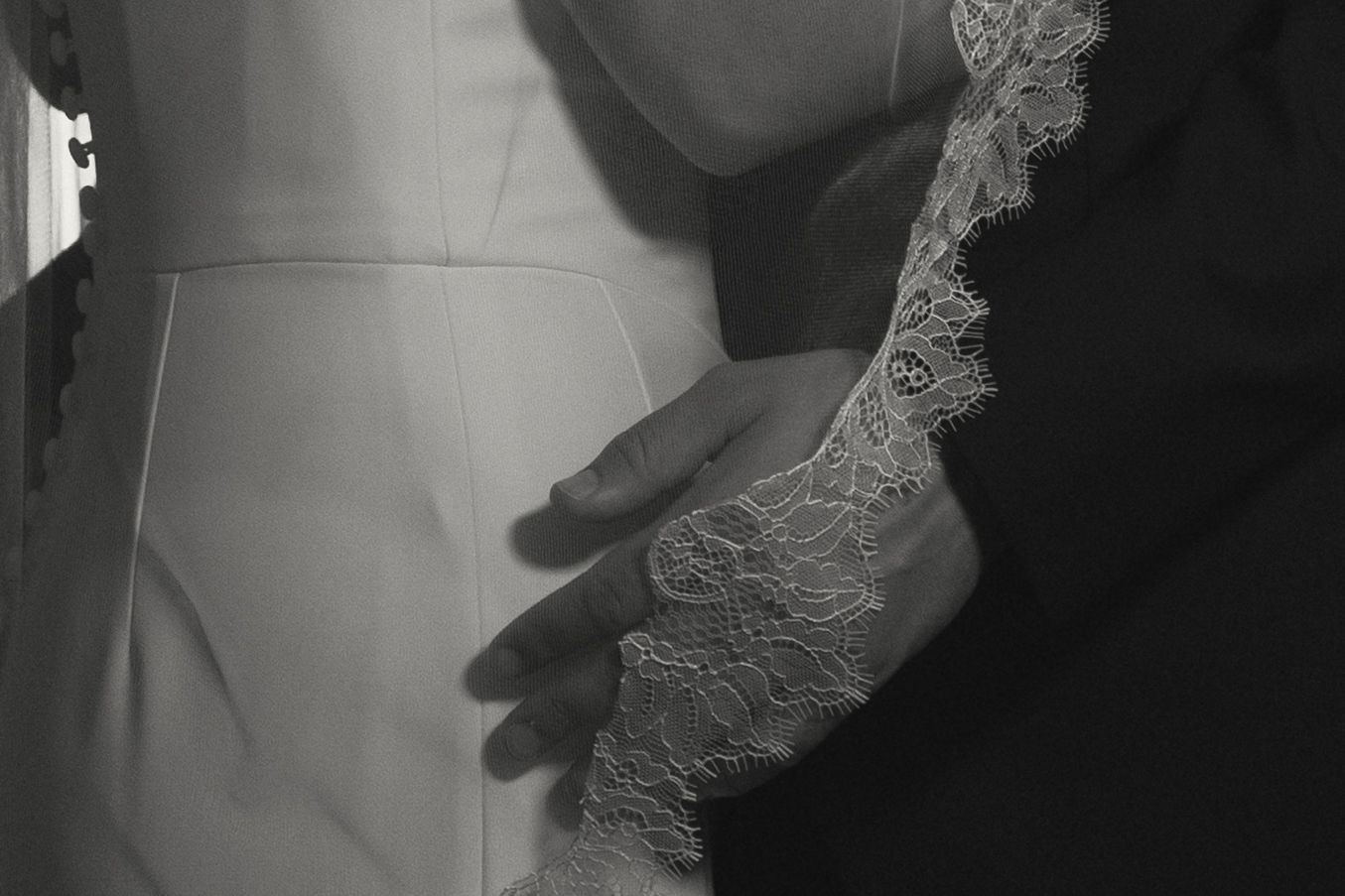 Close-up detail of delicate lace wedding dress and groom's hands in black and white.