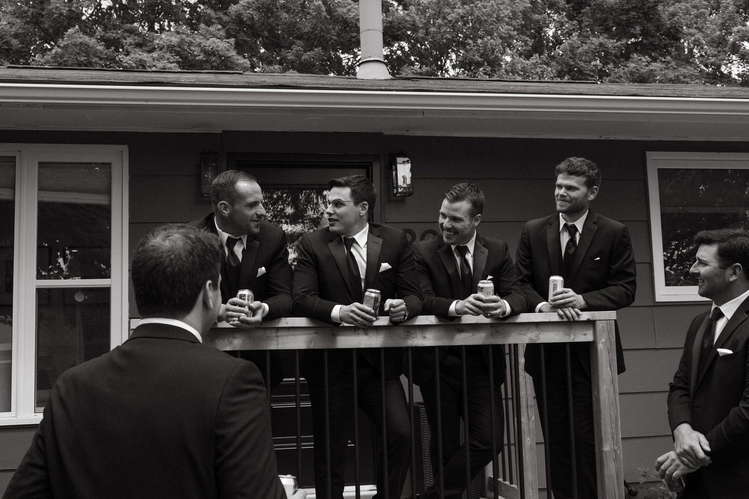 Wedding party members in dark suits gather outside a residential home.