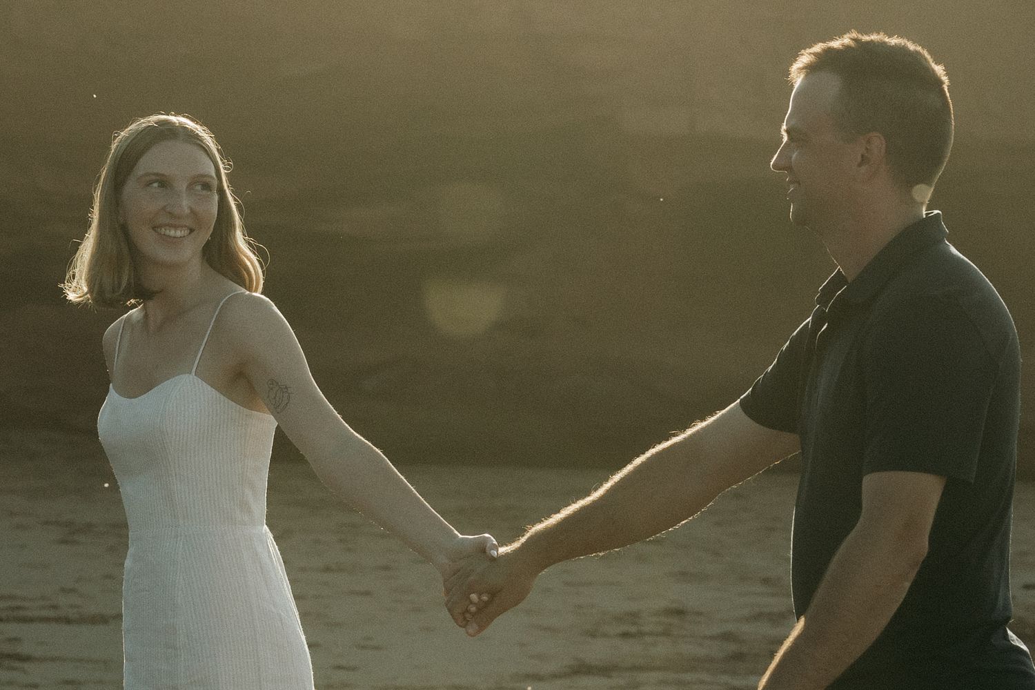 Couple holding hands at sunset on a beach with golden light.