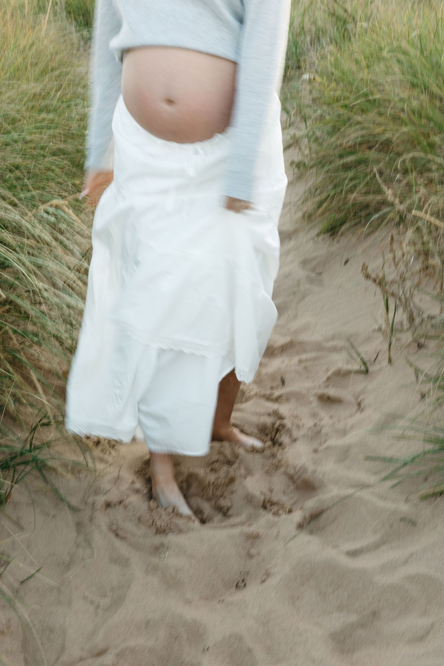 Close-up details of white maternity dress and bare belly against sandy beach setting.