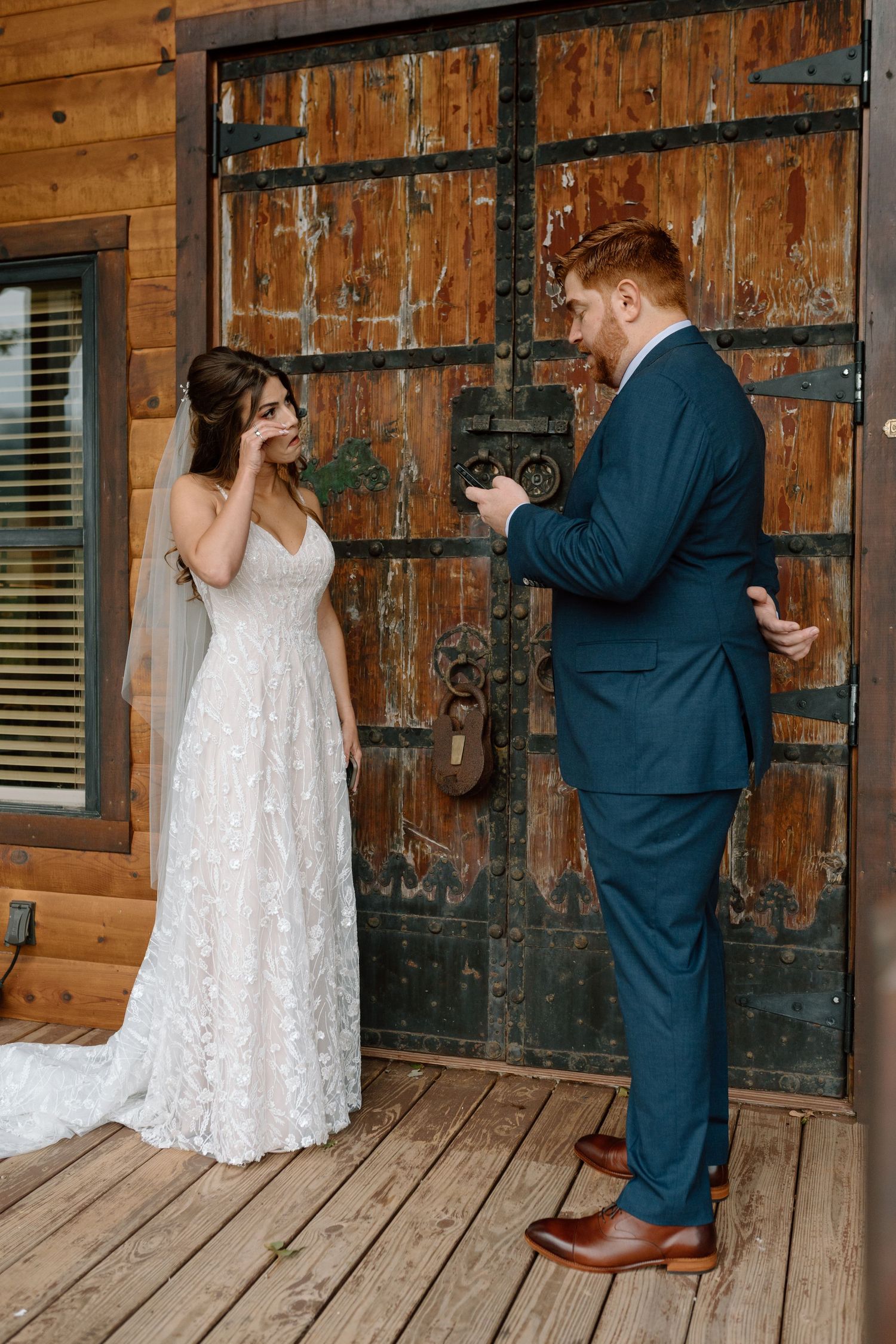 A couple in wedding attire stands in front of rustic wooden doors during their first look photo sequence.