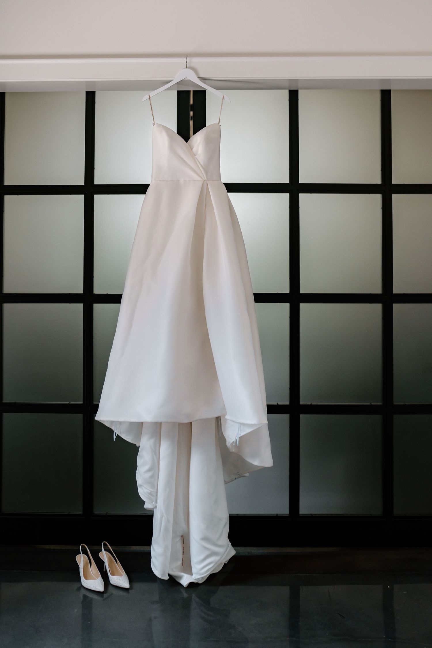 An elegant white wedding dress hangs on a hanger against a modern paneled wall with bridal shoes beneath.