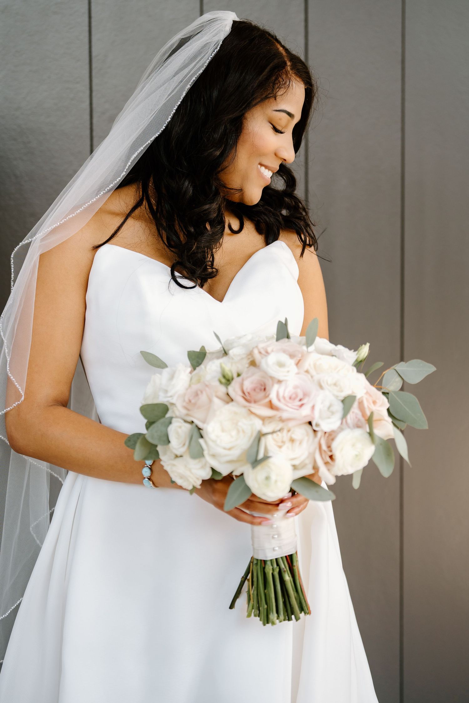 A bride in a white wedding dress holds a romantic bouquet of blush roses and eucalyptus against a gray wall.
