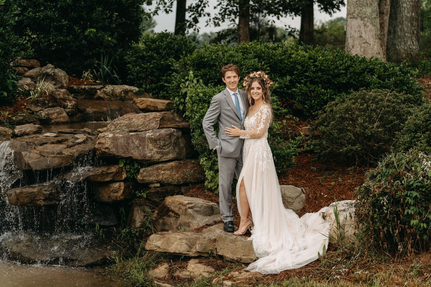 Bride and groom posing by a stone waterfall in a lush garden, bride wearing a floral crown and white gown.