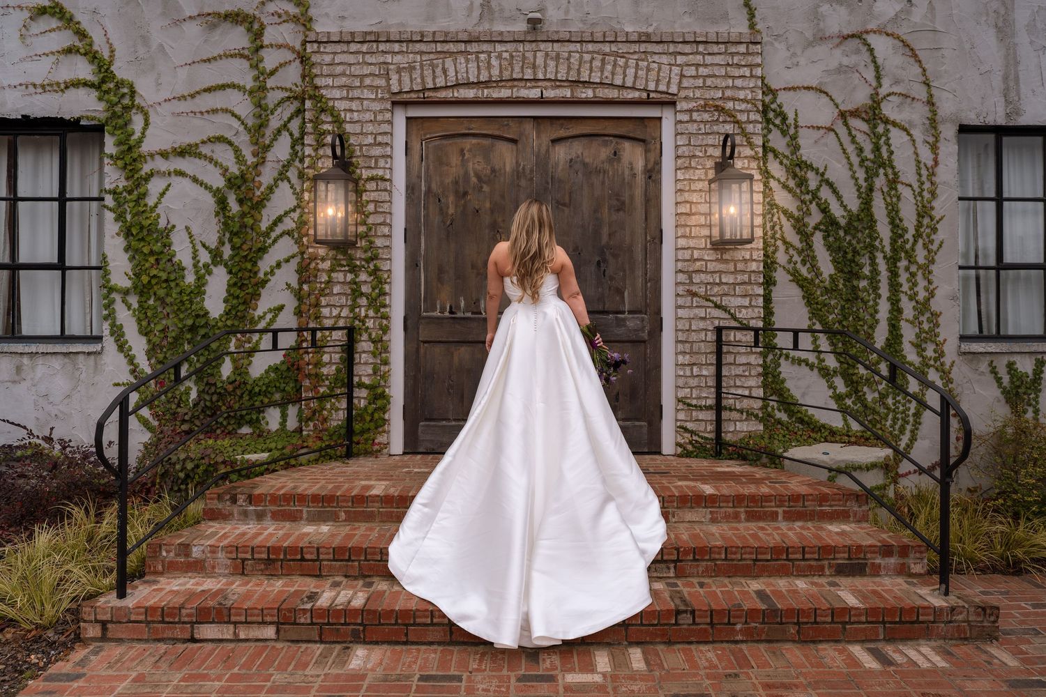 A bride in a flowing white wedding dress and cape stands in front of a rustic brick doorway.