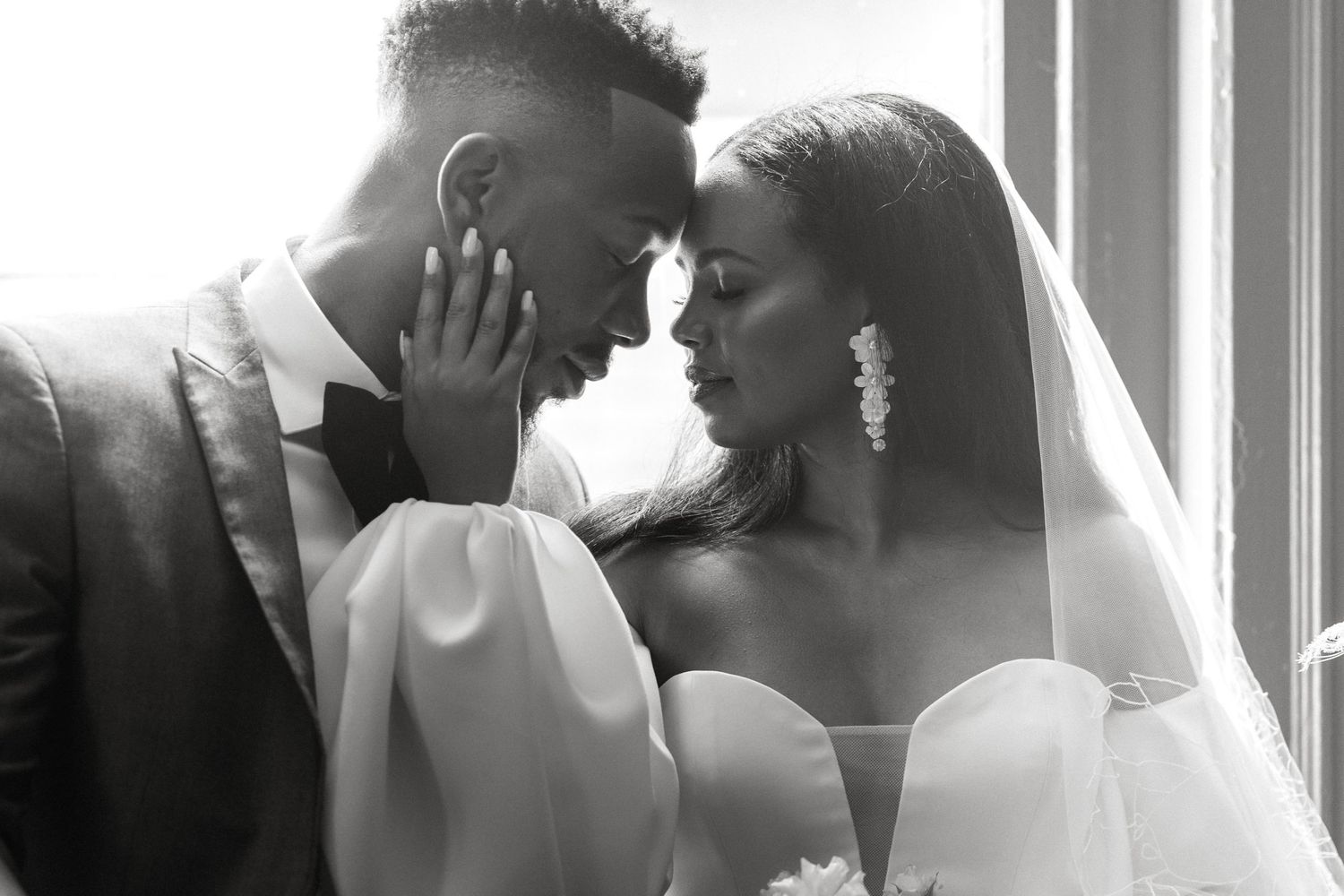A tender black and white wedding portrait shows a romantic moment between newlyweds by a sunlit window.
