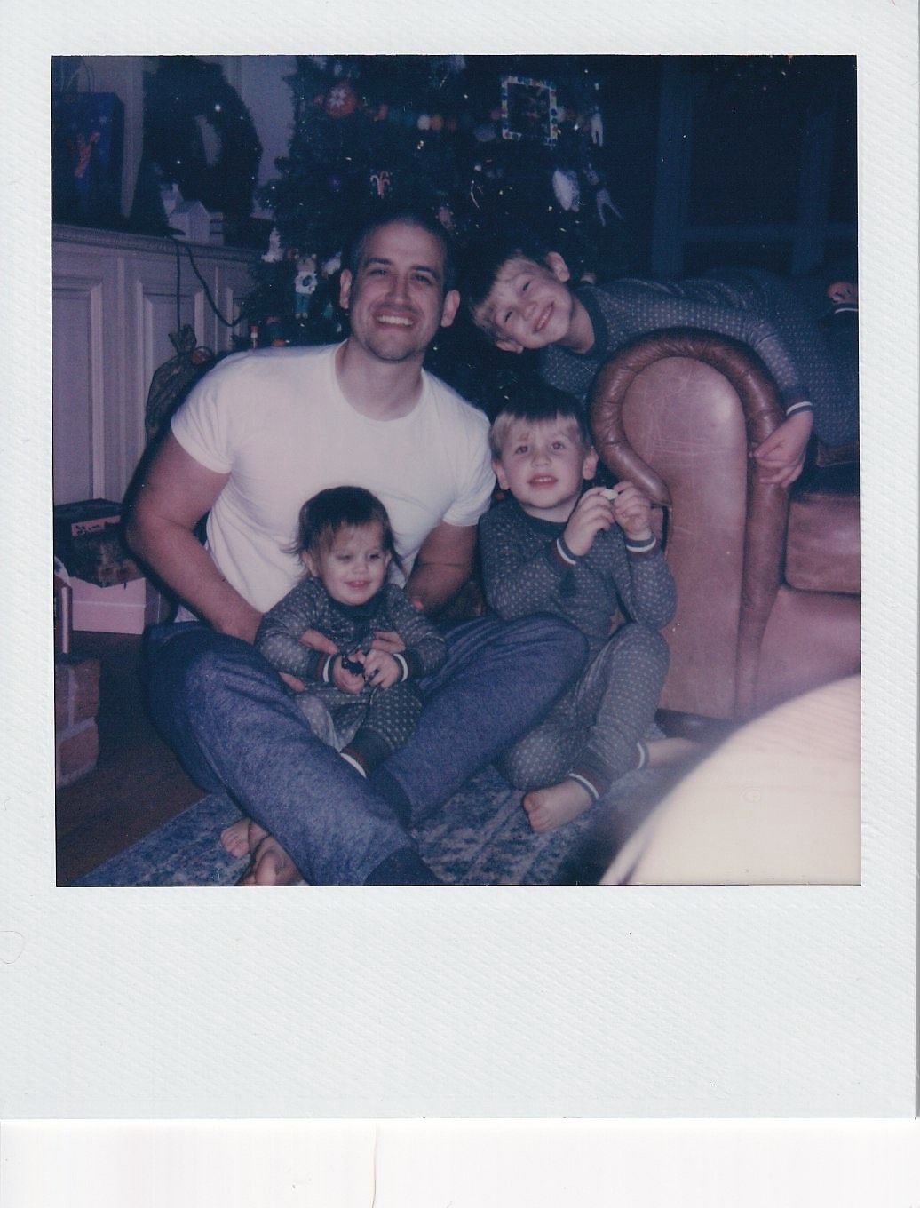 A man poses with three young children in front of a Christmas tree in a vintage Polaroid family photo.