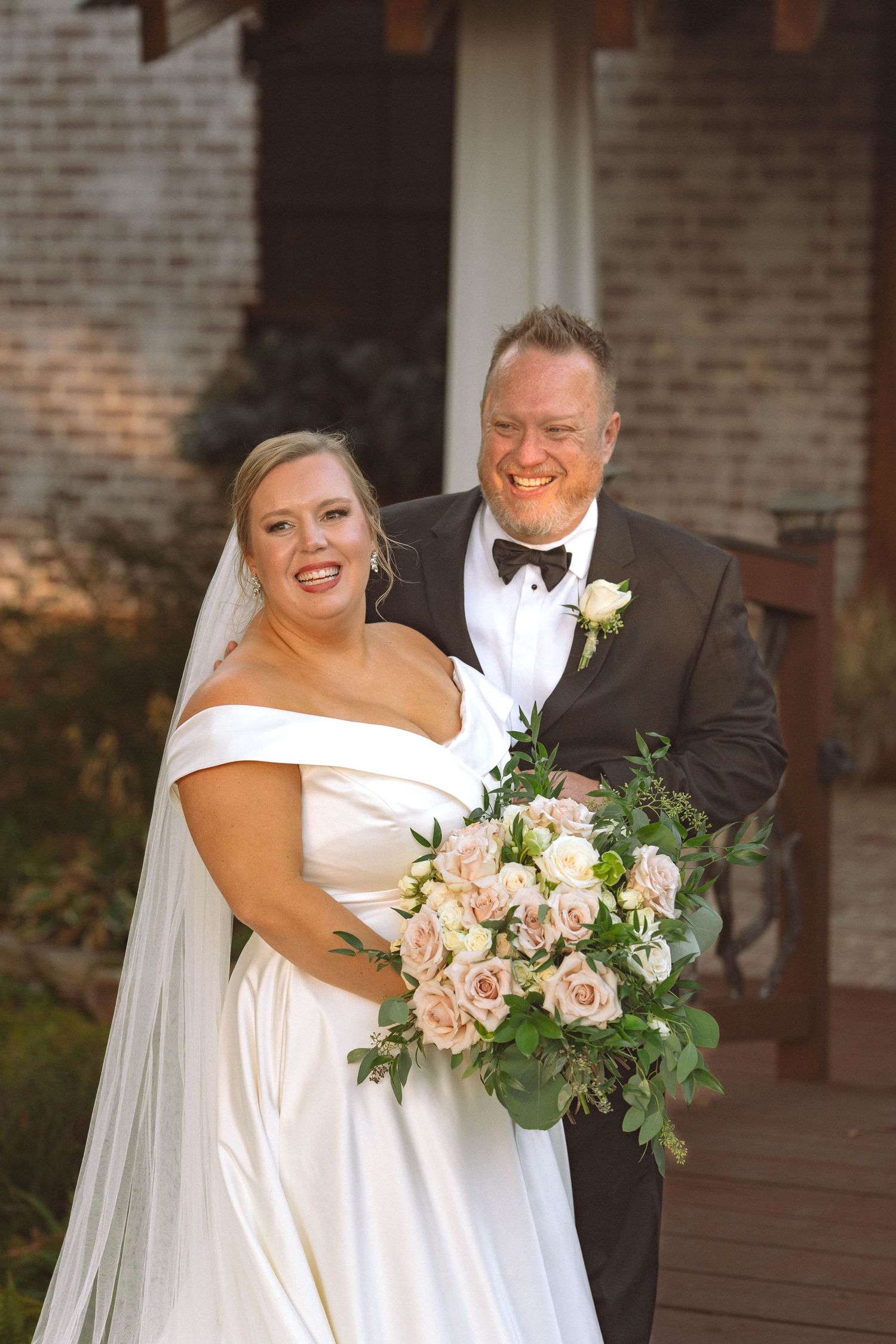 A bride in a white off-shoulder gown with a bouquet of pink roses poses with her companion against a brick backdrop.