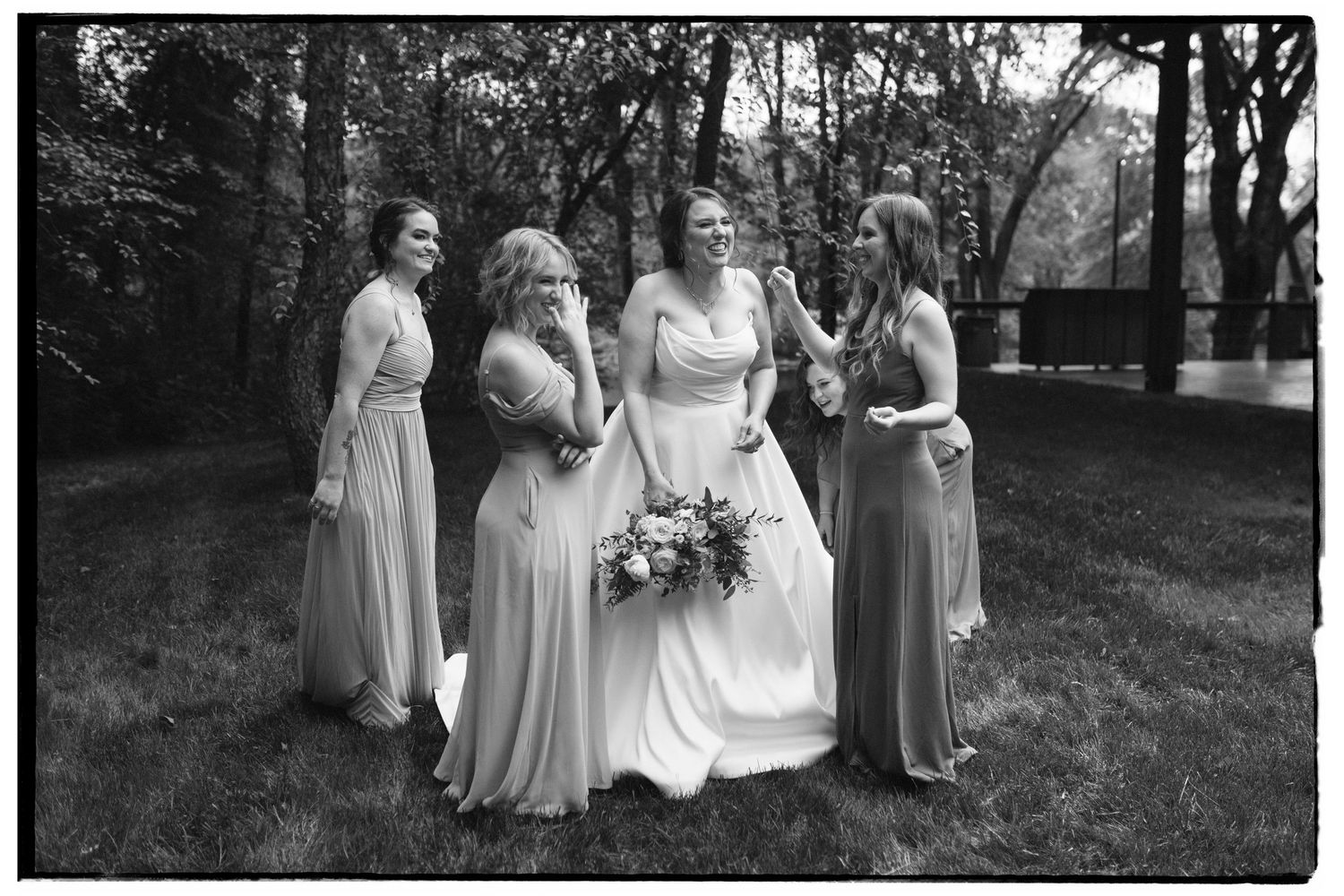 A bride in a white gown stands with three bridesmaids wearing matching dresses in a black and white outdoor photo.