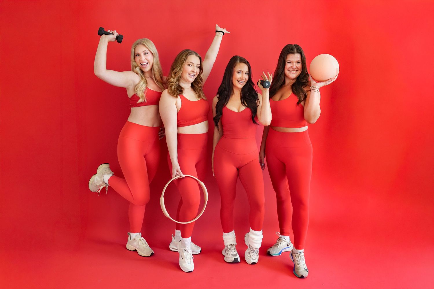 Four women in matching red athletic wear pose with fitness equipment against a vibrant red background.