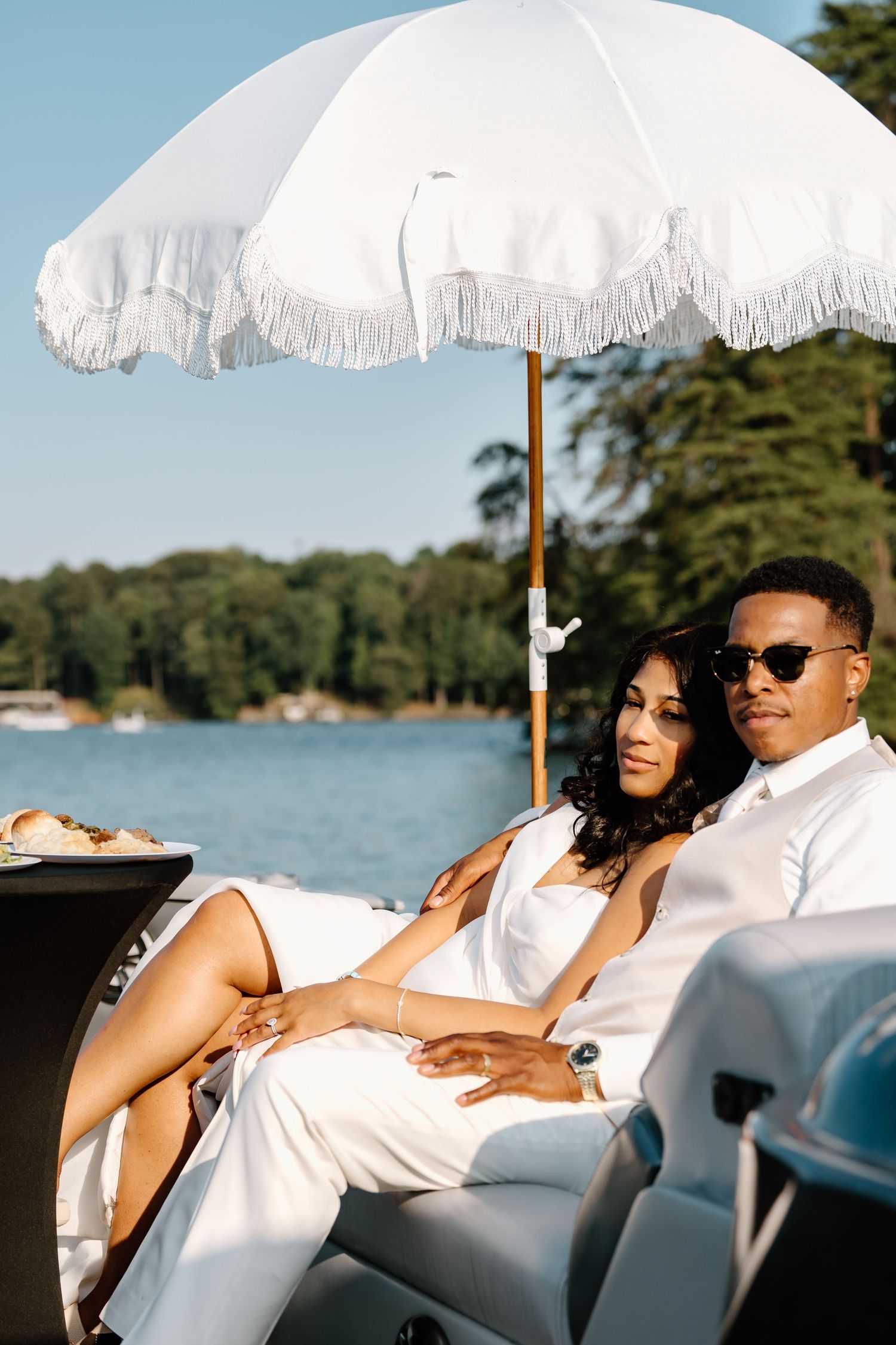 Elegant couple relaxes on white boat under vintage fringe umbrella by scenic lake on sunny summer day.