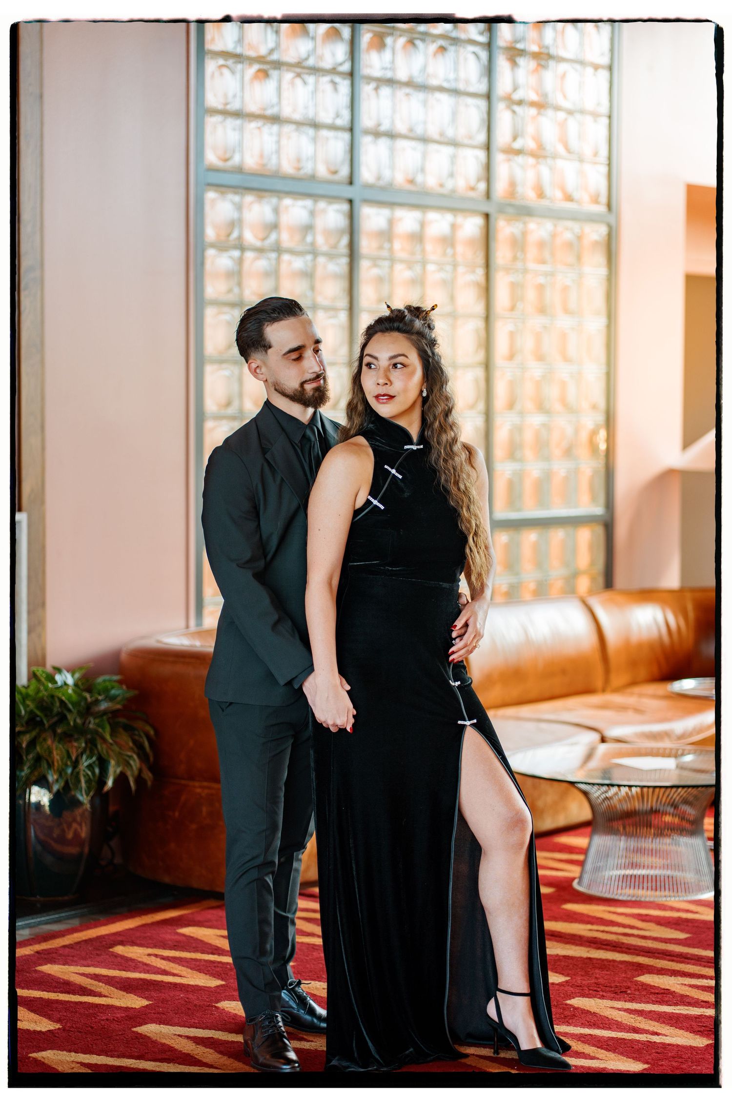 A couple in formal attire poses together in an elegant venue with glass block windows and red carpet.