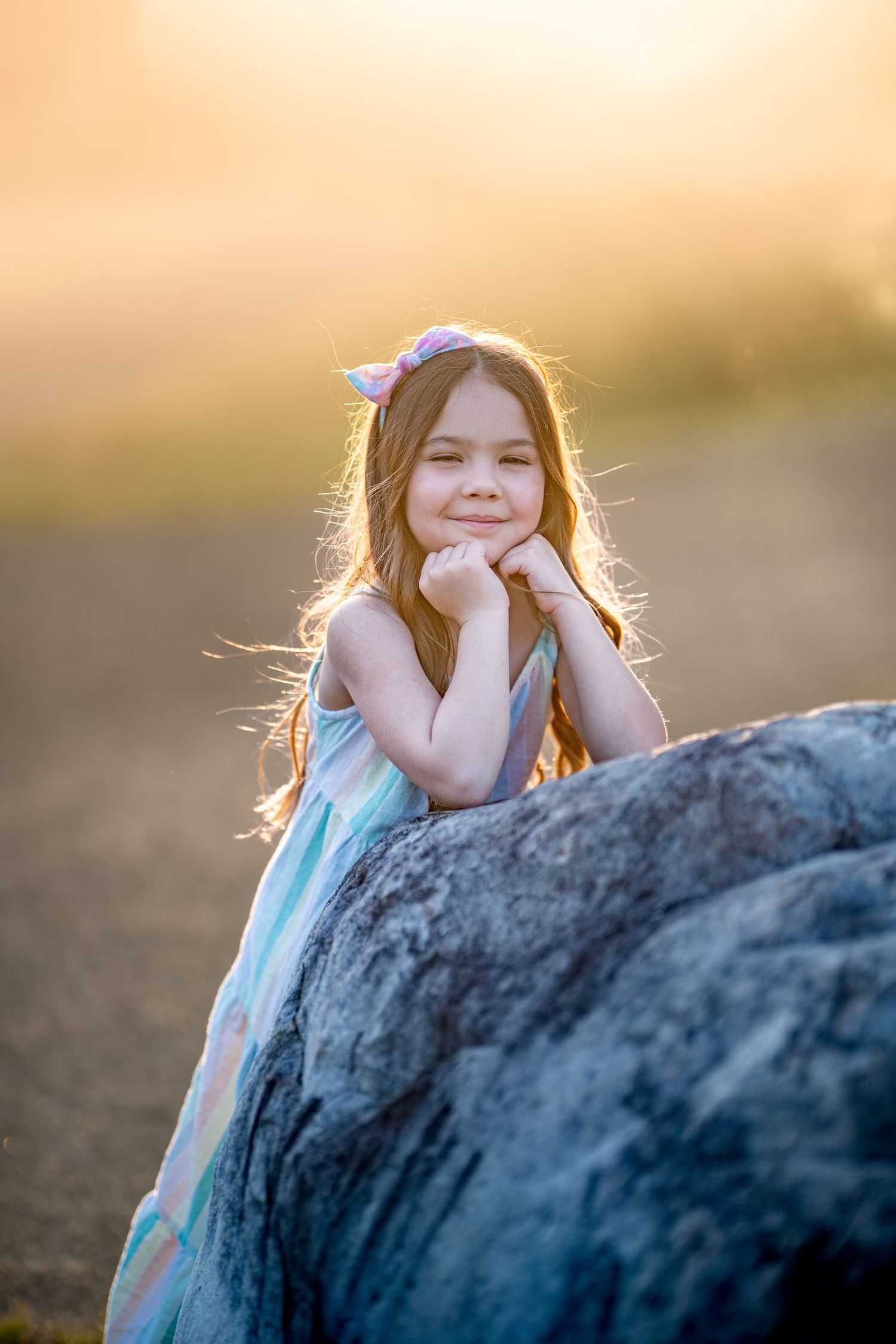 A child in a blue dress sits on a rock at sunset with golden light creating a dreamy backlit effect.