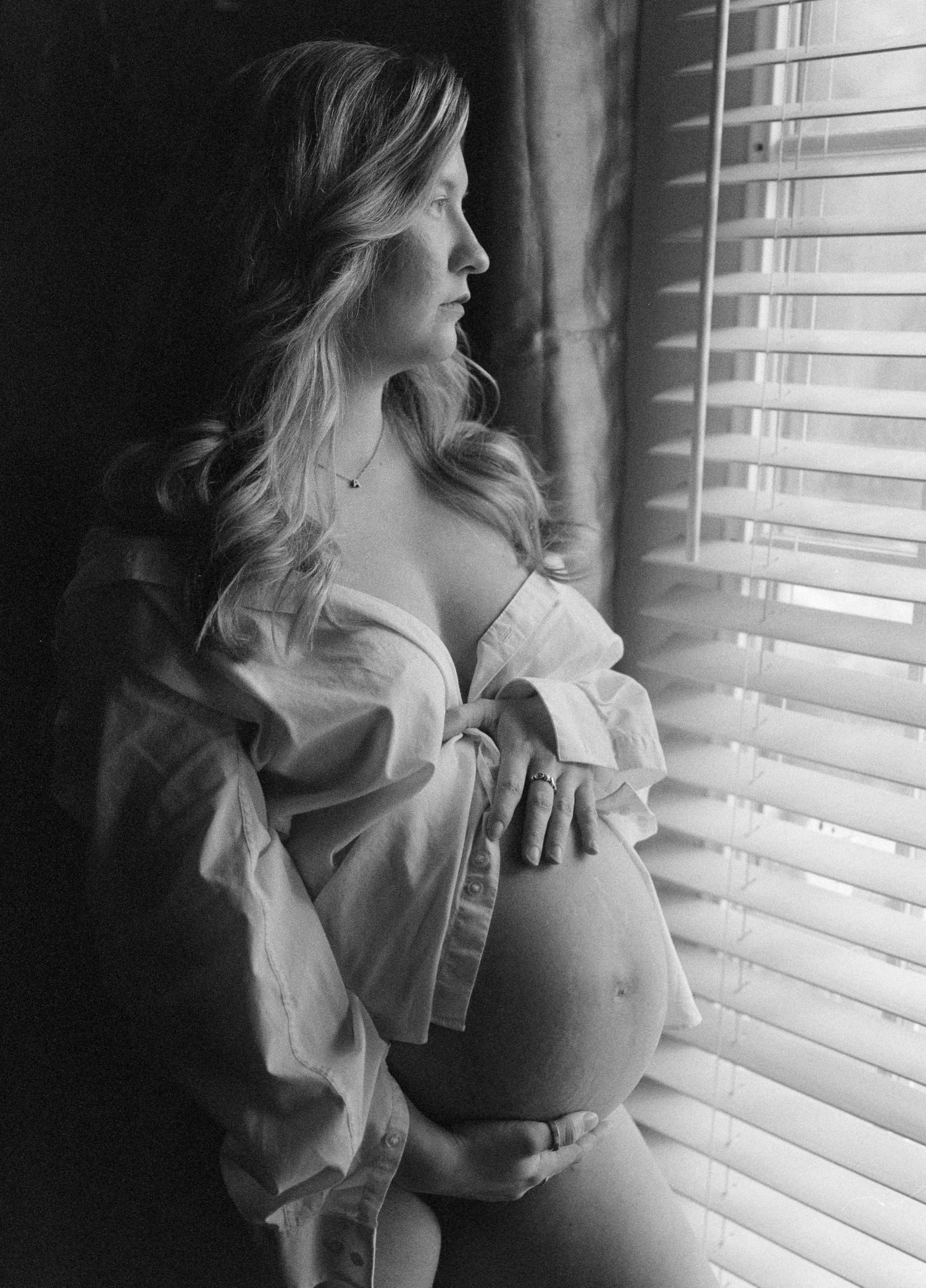 Artistic black and white maternity photos showing an expectant mother by window blinds in soft natural light.