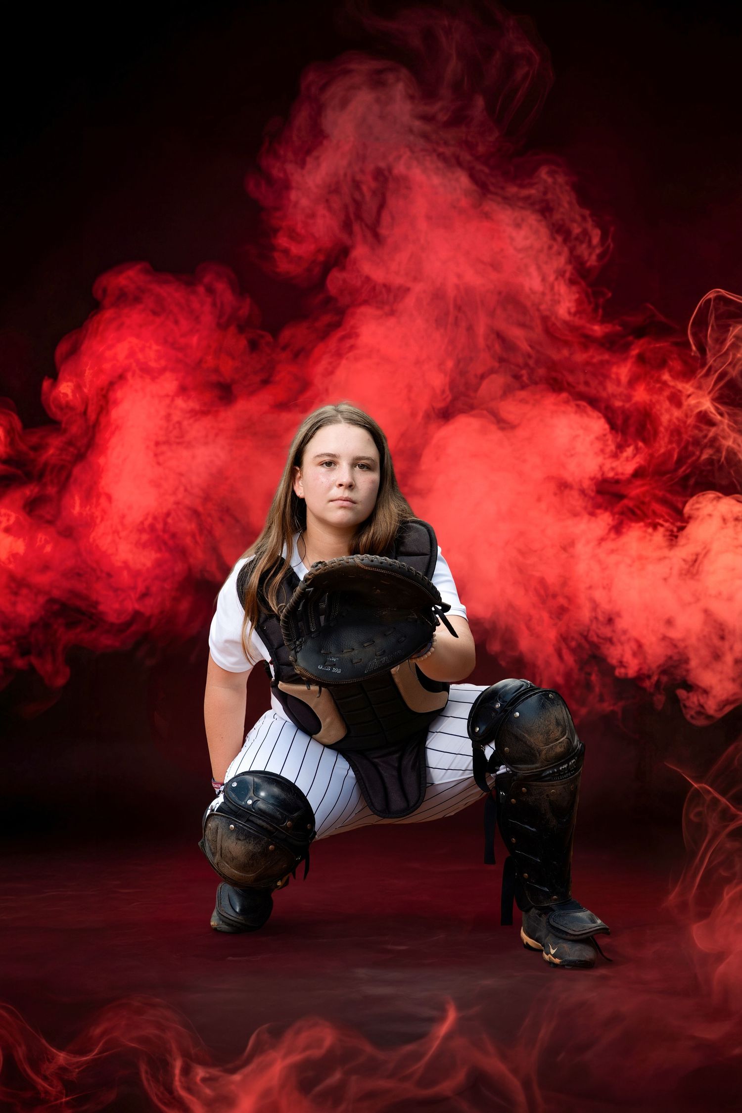 Female softball catcher in uniform crouching with mitt, surrounded by dramatic red smoke on dark background.