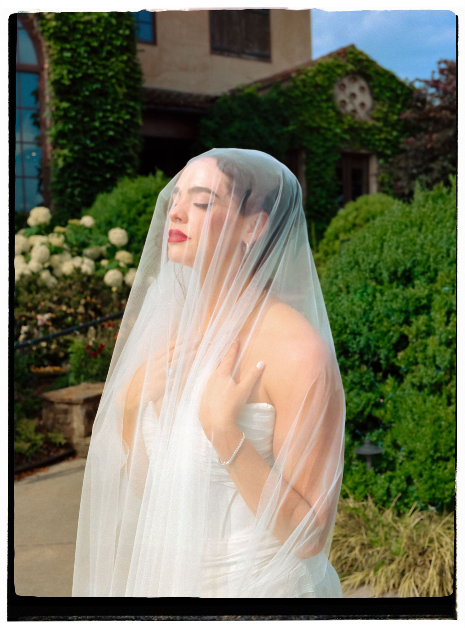 A bride in a flowing white veil poses against a lush garden backdrop in a romantic outdoor setting.
