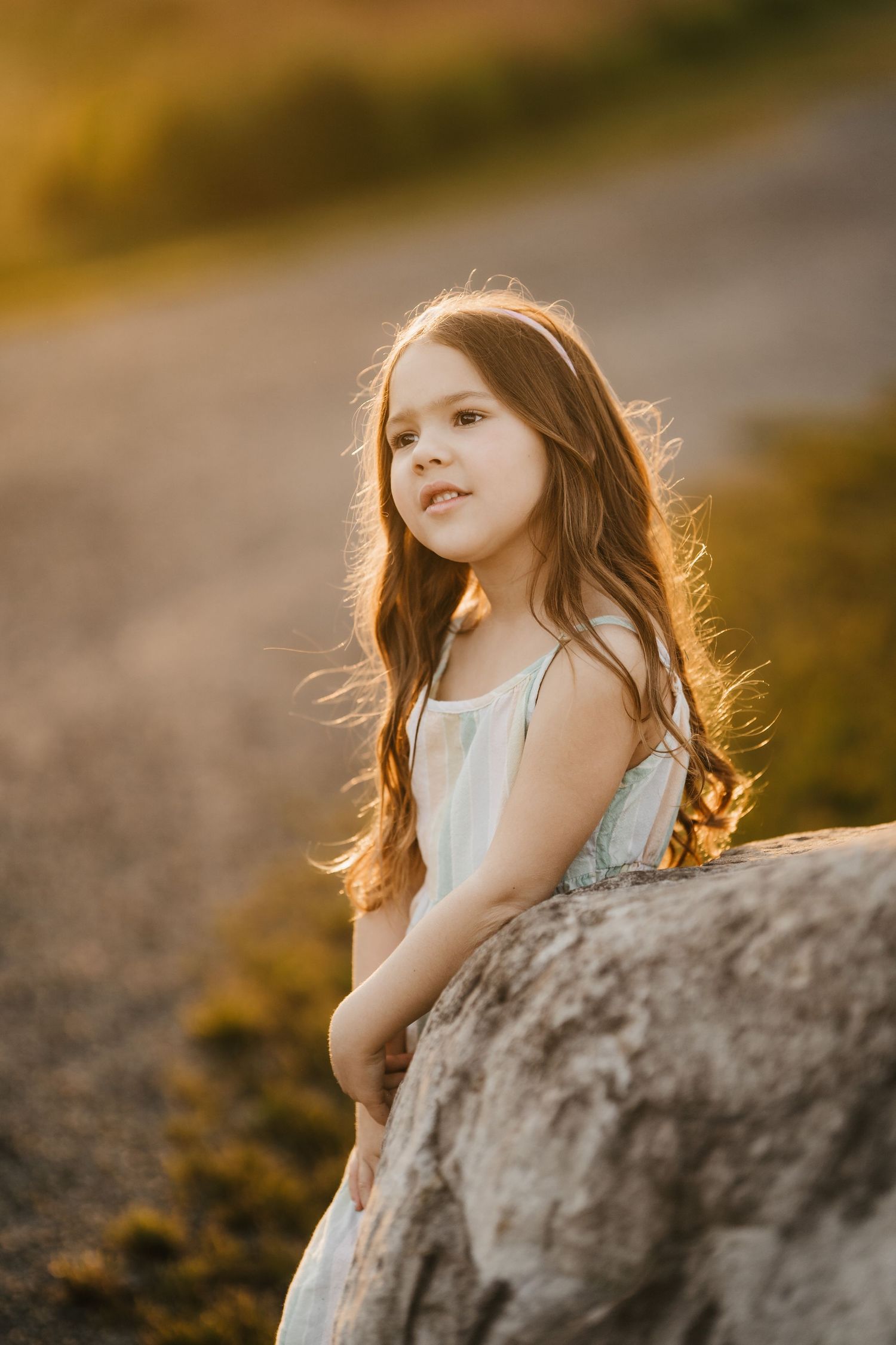 A child in a white dress poses near a rock formation during golden hour with sunlit hair flowing in the breeze.