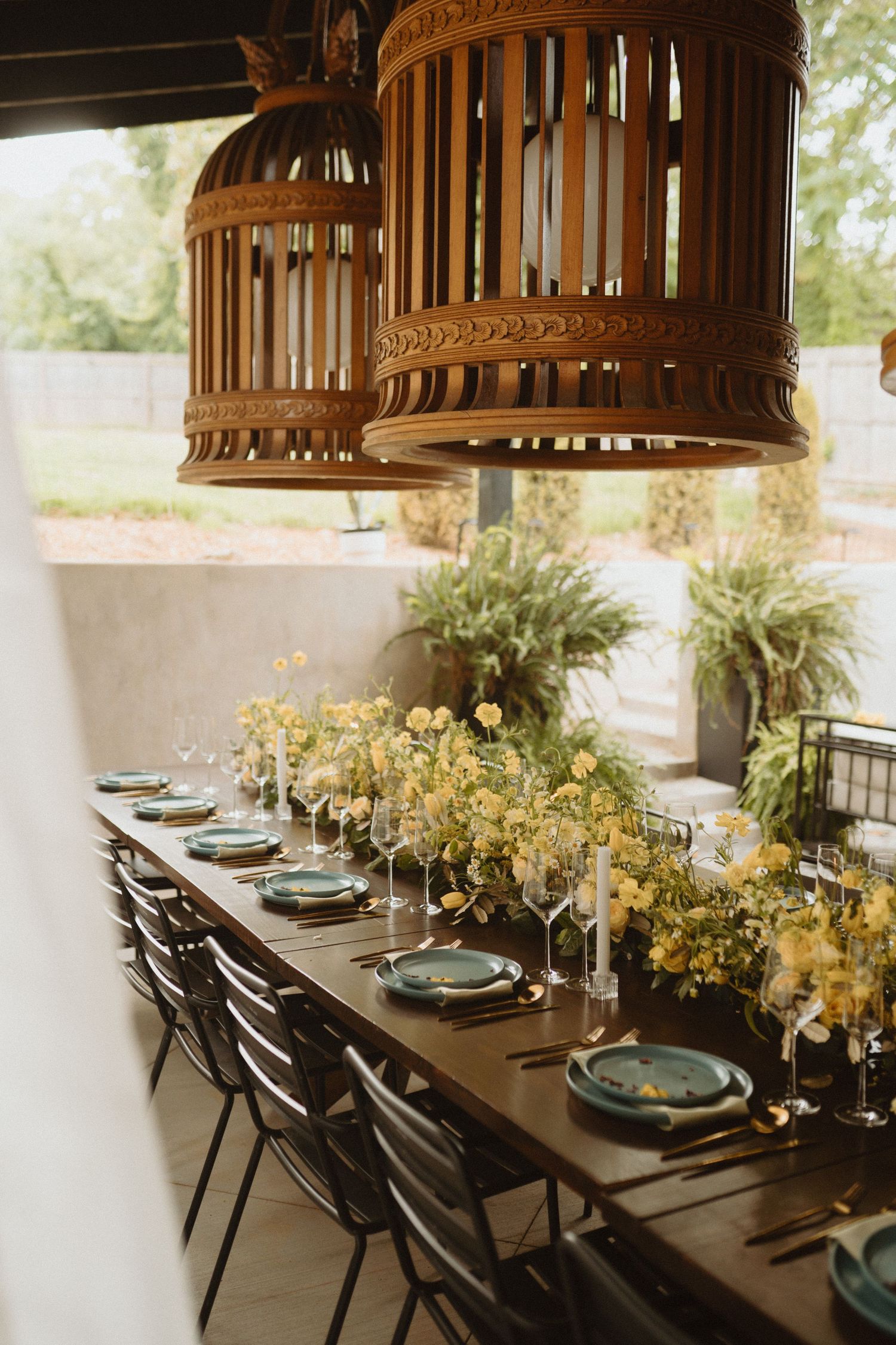 Elegant dining table setup with dark wood, green plates, and woven pendant lights decorated with yellow floral arrangements.