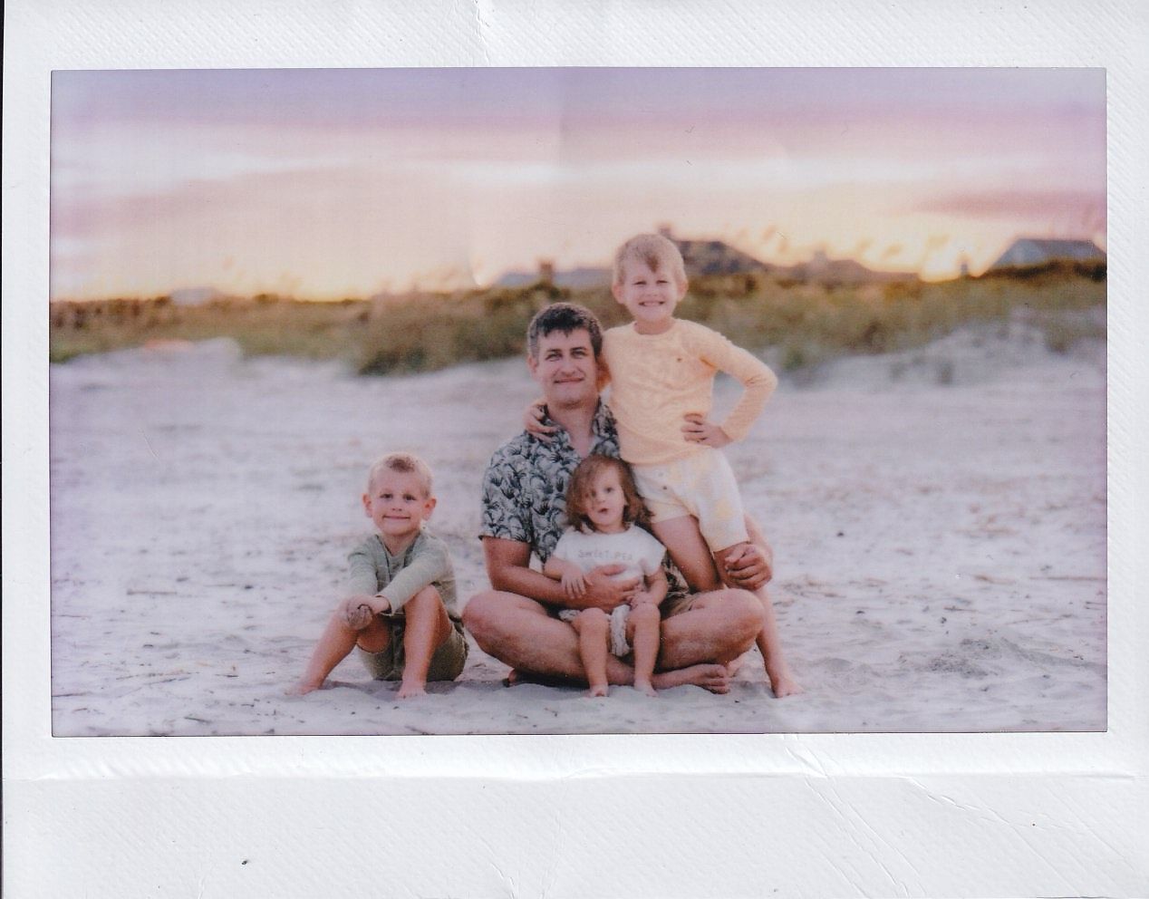 Family of four poses on sandy beach at sunset, father sitting with three young children smiling for a Polaroid-style photo.