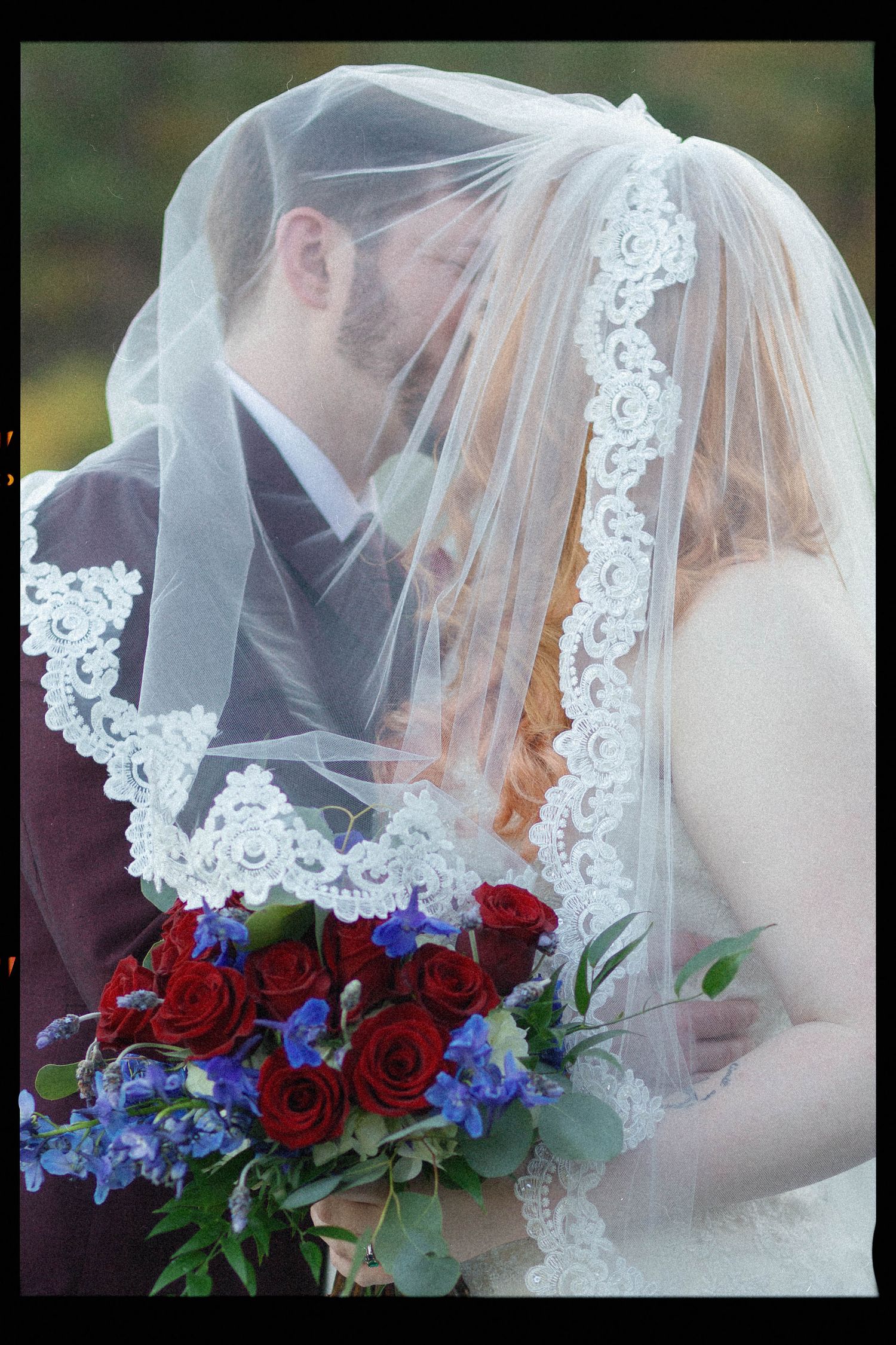 A romantic wedding moment captured through delicate lace veil with vibrant red roses and blue delphinium bouquet.