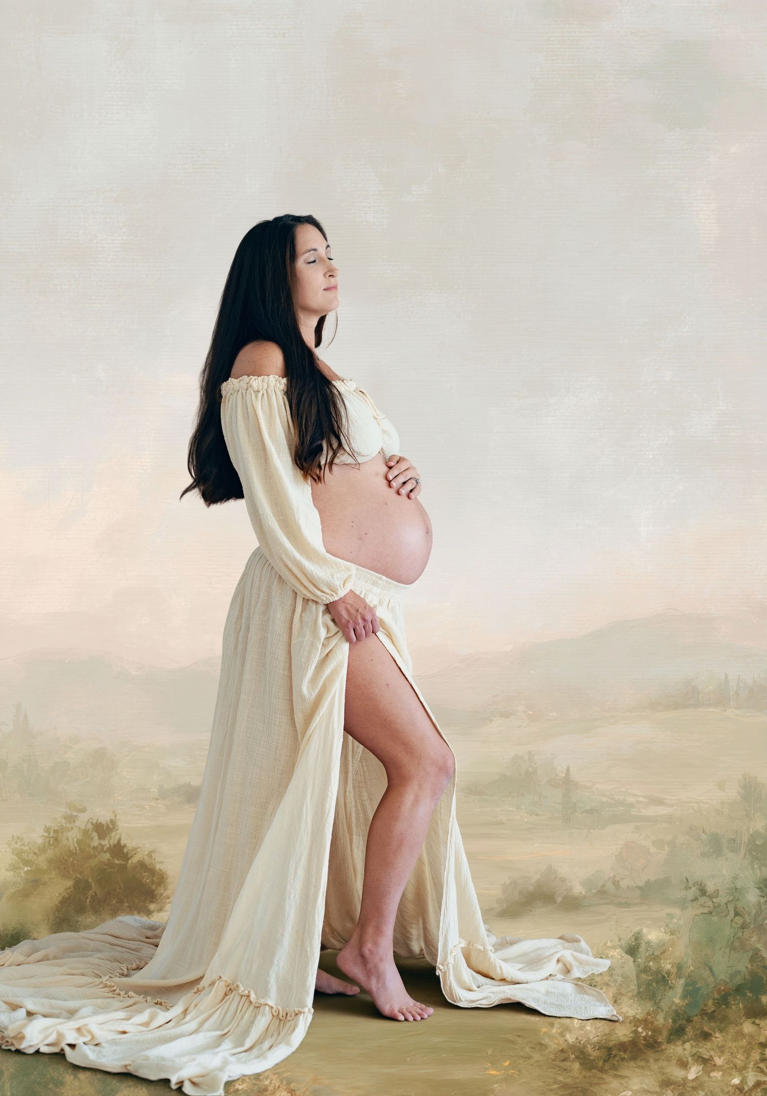 Pregnant woman in flowing cream off-shoulder gown cradling belly against soft pastoral painted backdrop.