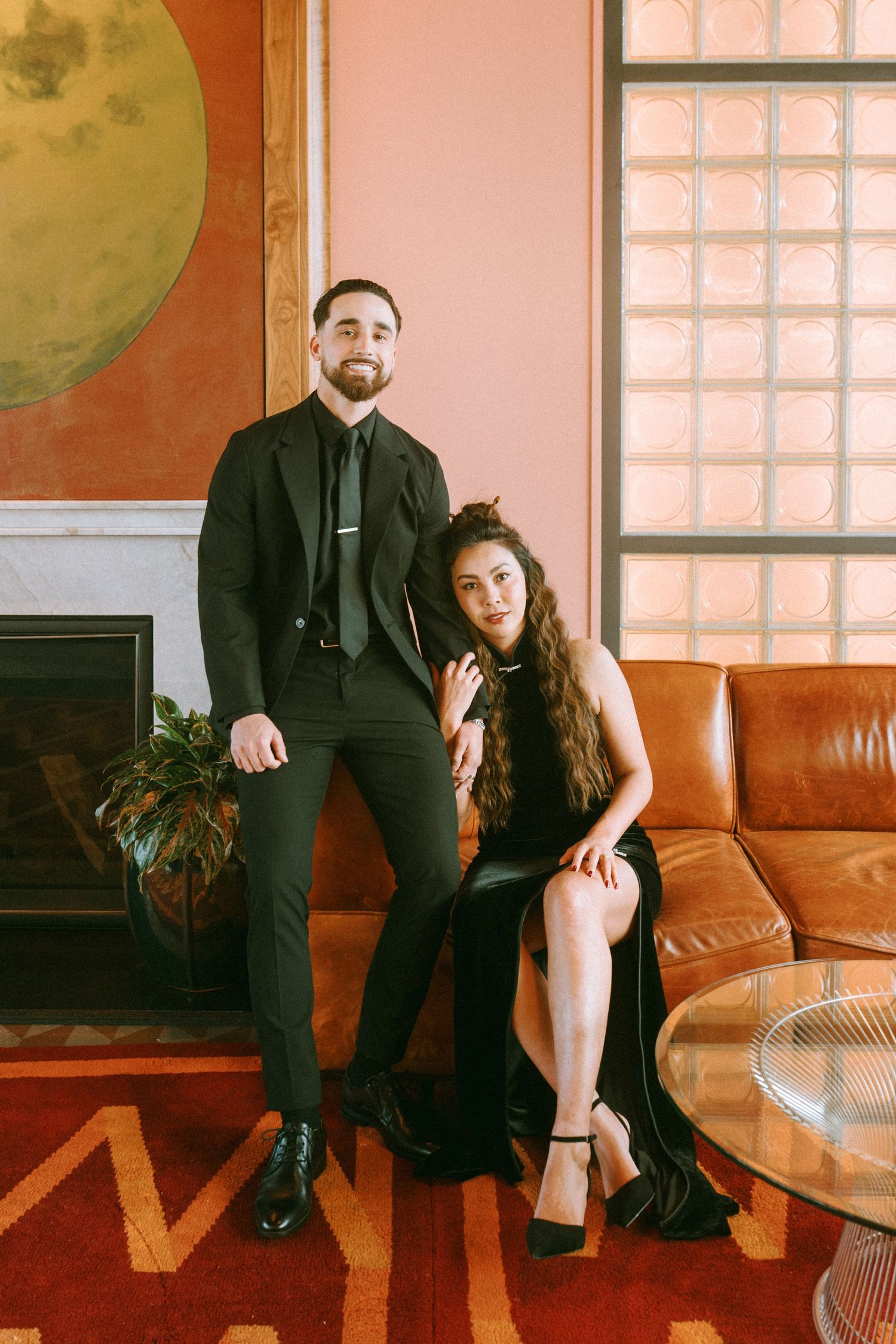 Two people in black formal attire sit together on a brown leather couch in a warm-lit room with glass block windows.