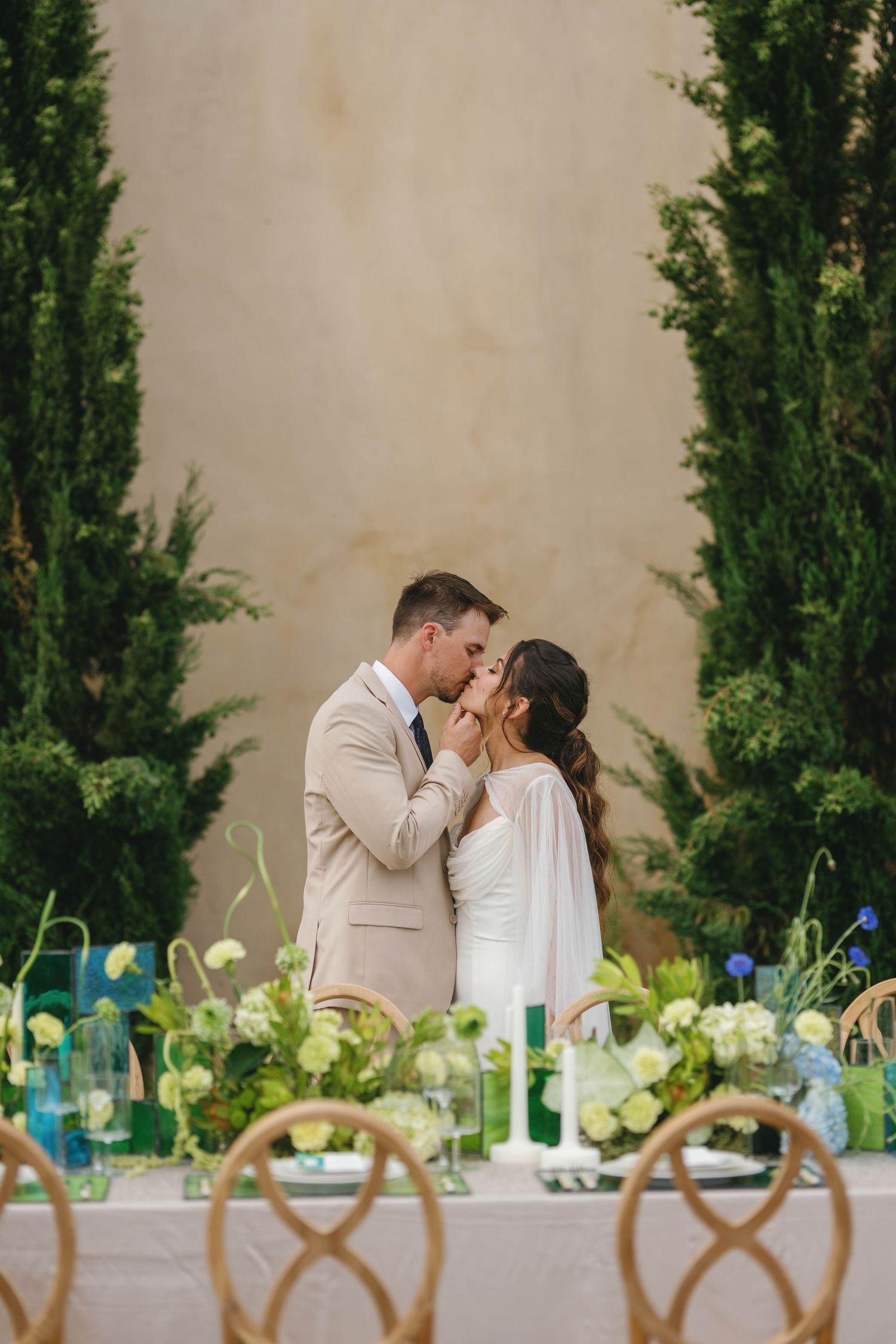 Romantic outdoor wedding reception table setting with elegant floral centerpieces and cypress trees in background.