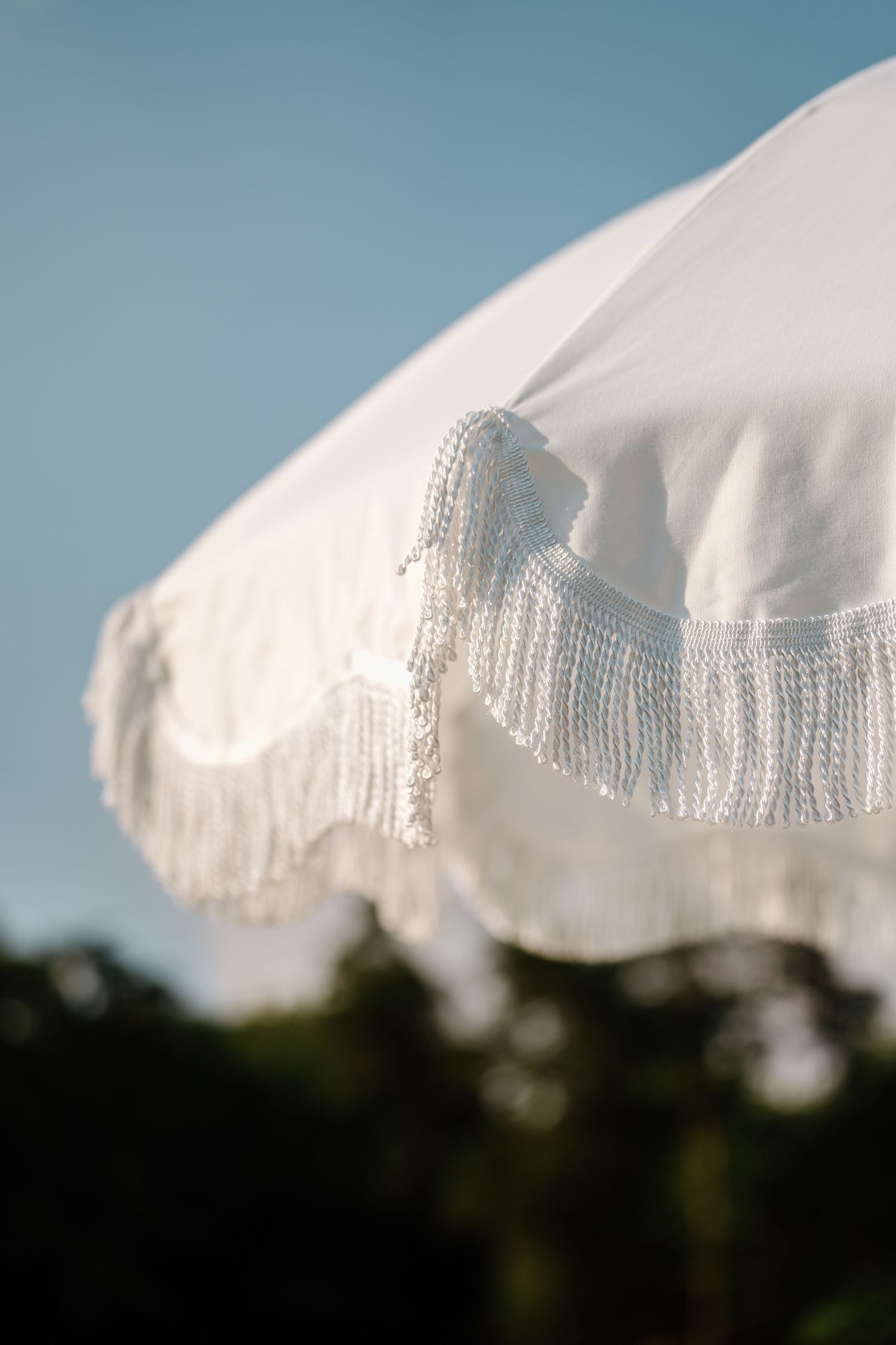 White fabric with delicate fringe trim billows against a blue sky with trees in the background.
