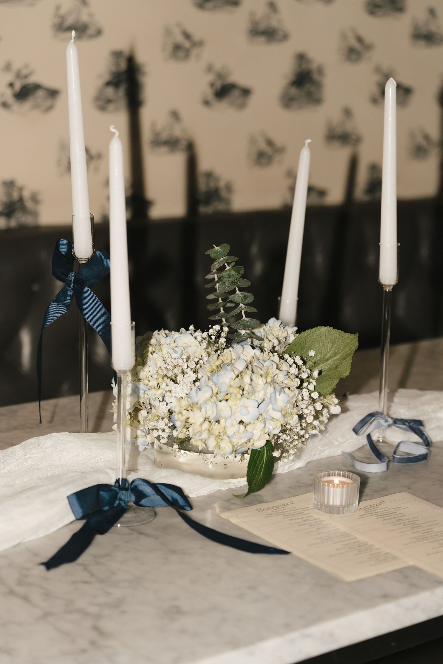 Elegant wedding table with white taper candles, blue ribbons, hydrangea centerpiece, and open book on marble surface.
