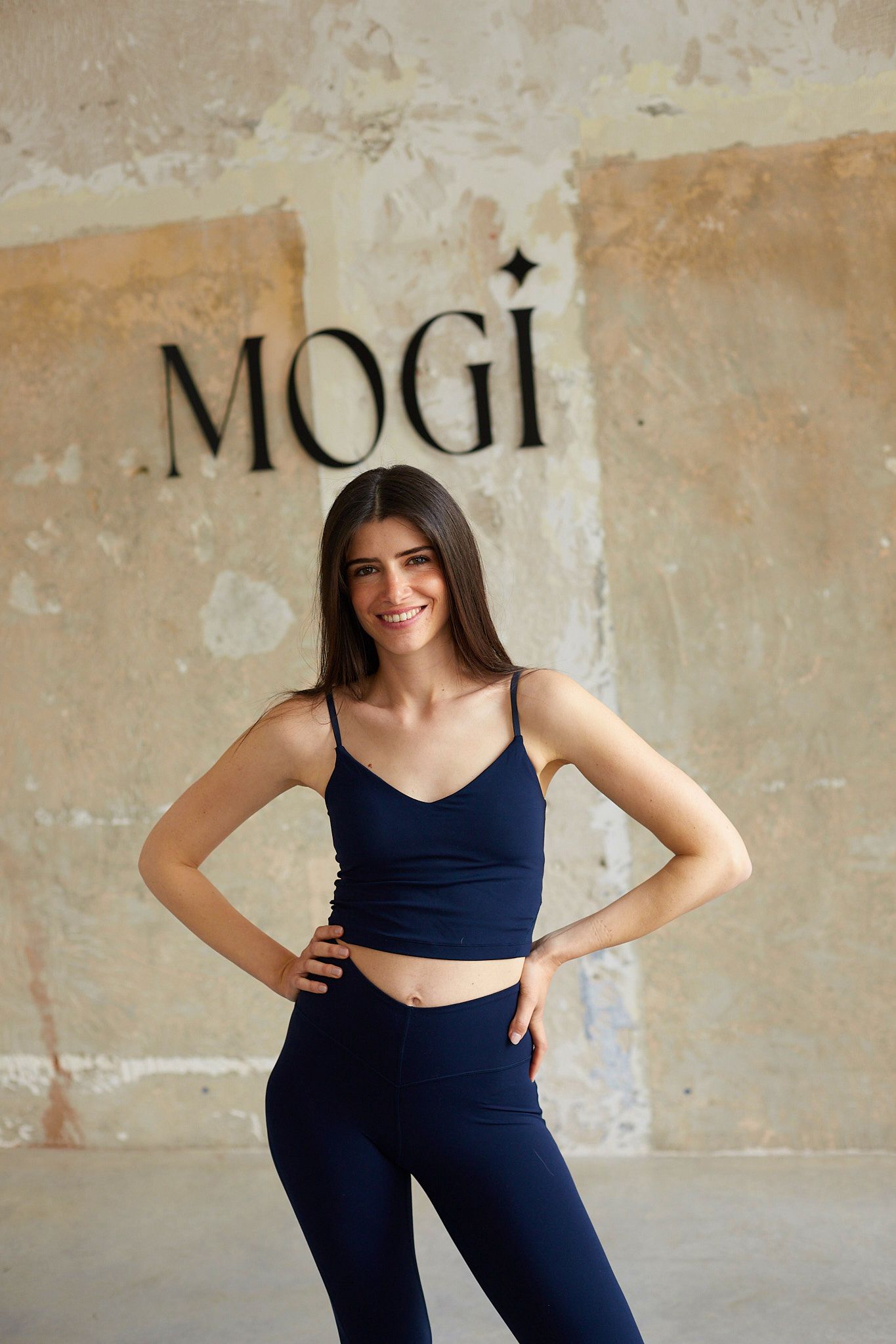 Woman in navy workout set poses confidently against rustic wall with MOGI signage.