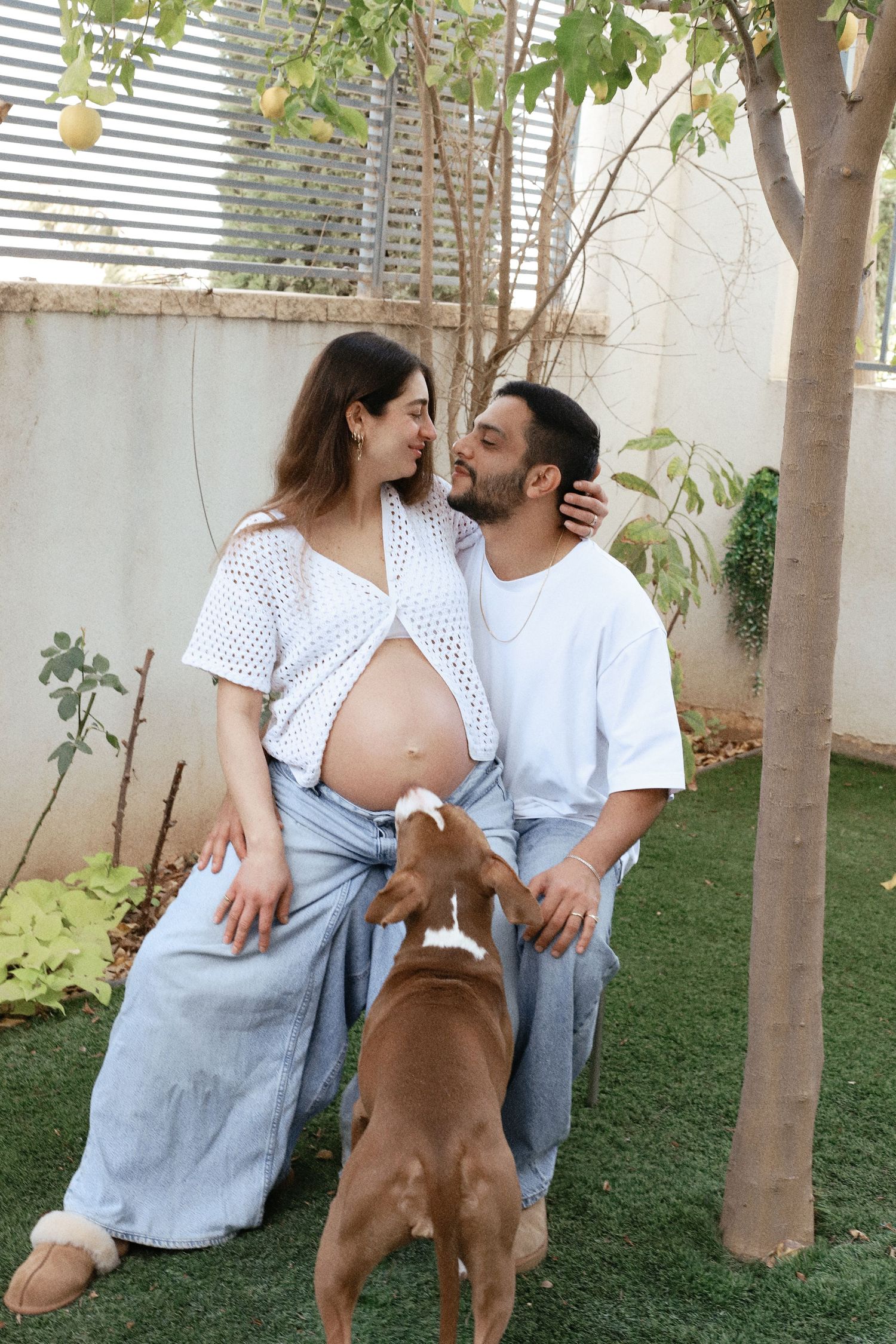 Expecting couple with dog share tender moment in backyard garden setting.