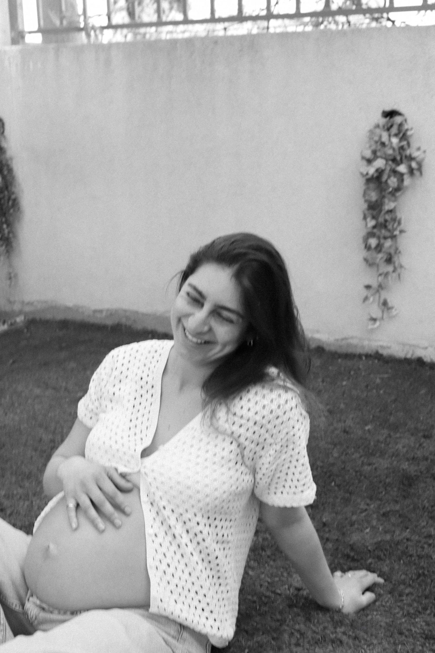 Expectant mother in white crochet top smiles while sitting outdoors in black and white photo.