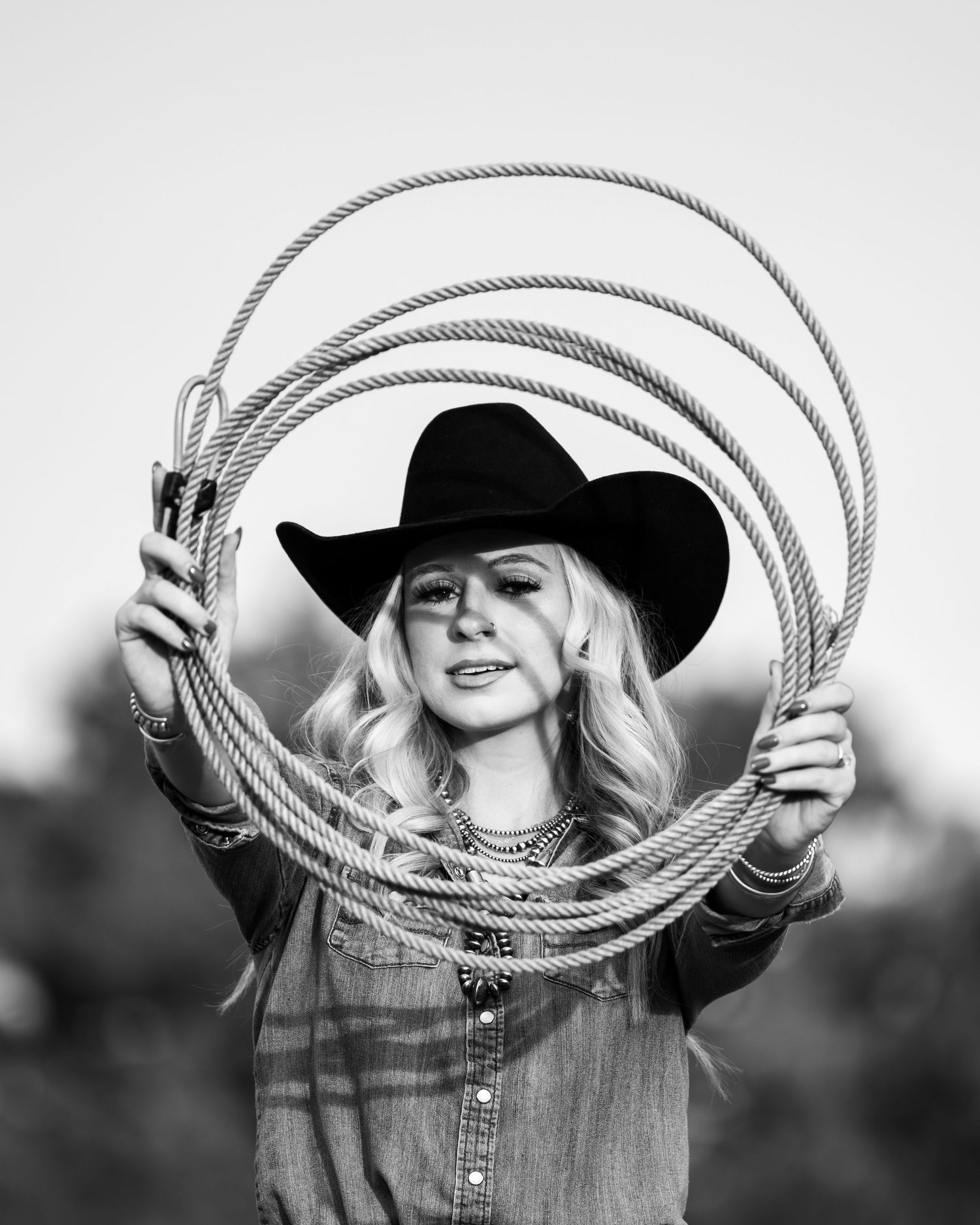 Western-styled figure in denim and black cowboy hat holds up a lasso rope in black and white portrait.