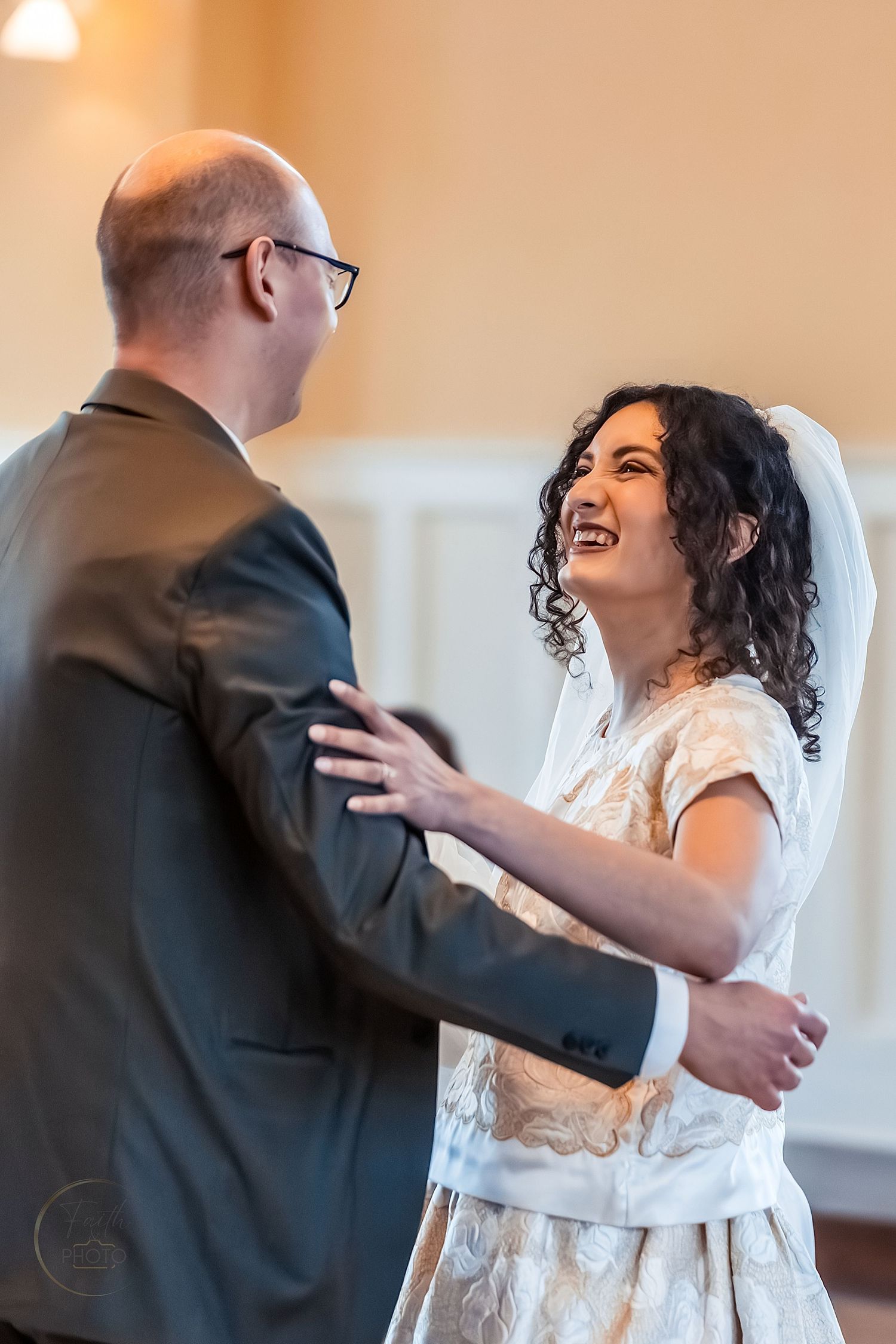 A bride in a white lace dress laughs joyfully while sharing a first dance with her groom in a warmly lit venue.